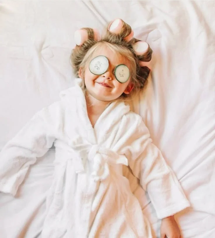 Une jeune fille allongée dans un lit, portant une robe de chambre blanche, avec des rondelles de concombre sur les yeux, entourée de doigts en forme de oreilles sur sa tête, souriant.