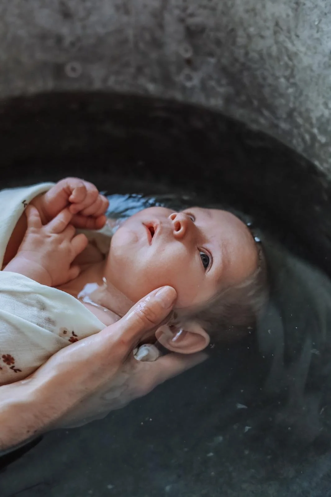 Bébé en train d'être baptisé ou de recevoir un rituel dans l'eau, avec des mains soutenant sa tête et son corps dans une cuve ou un bassin en pierre.