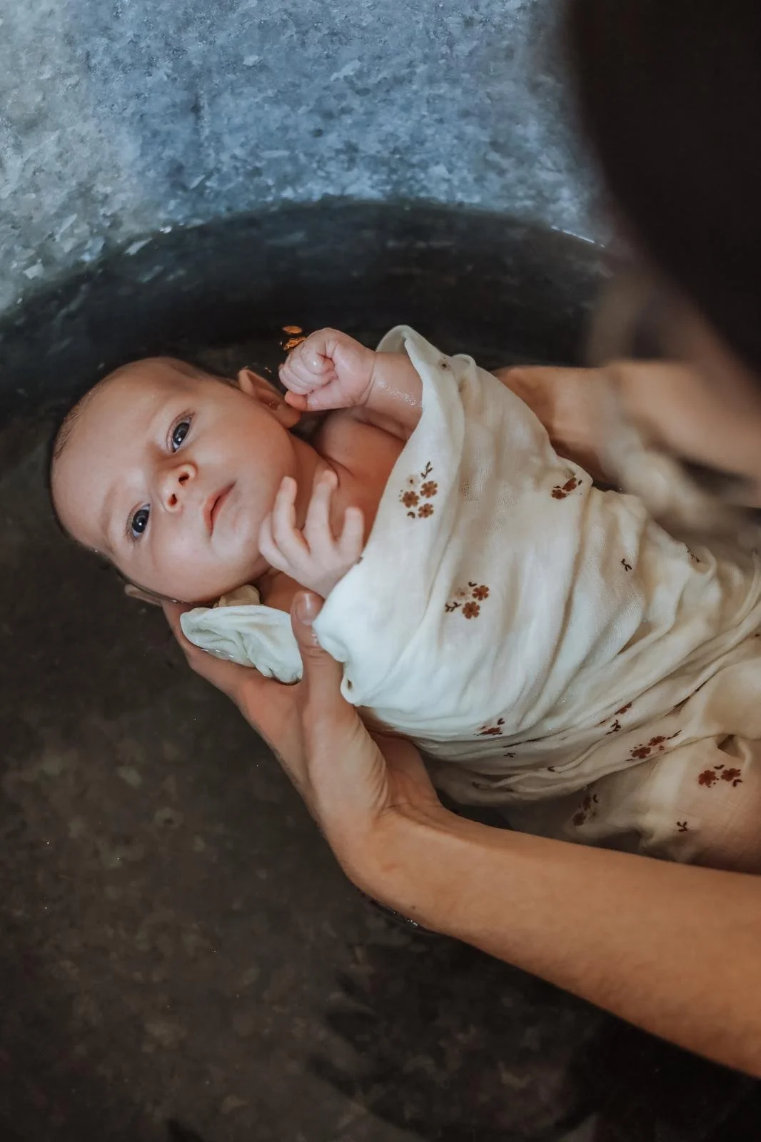 Un bébé lavé dans l'eau, entouré d'une main aidante, portant une tenue beige à petites fleurs marron.