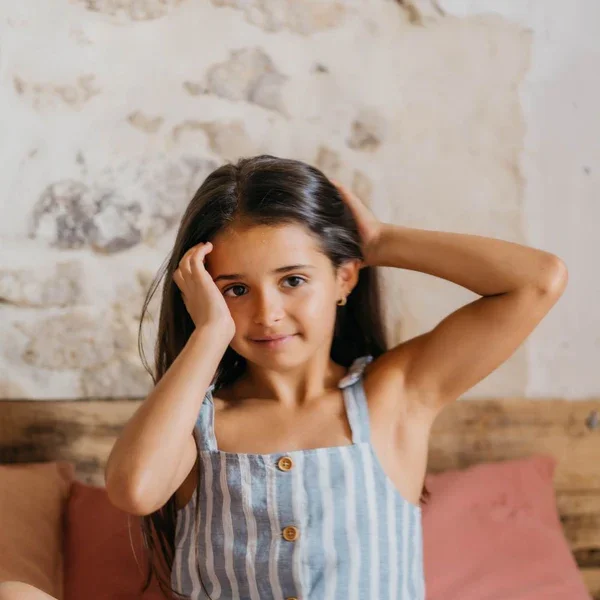 Une jeune fille souriante aux cheveux longs pose avec une main derrière la tête dans une chambre avec un mur en pierre et un lit avec des oreillers rose.
