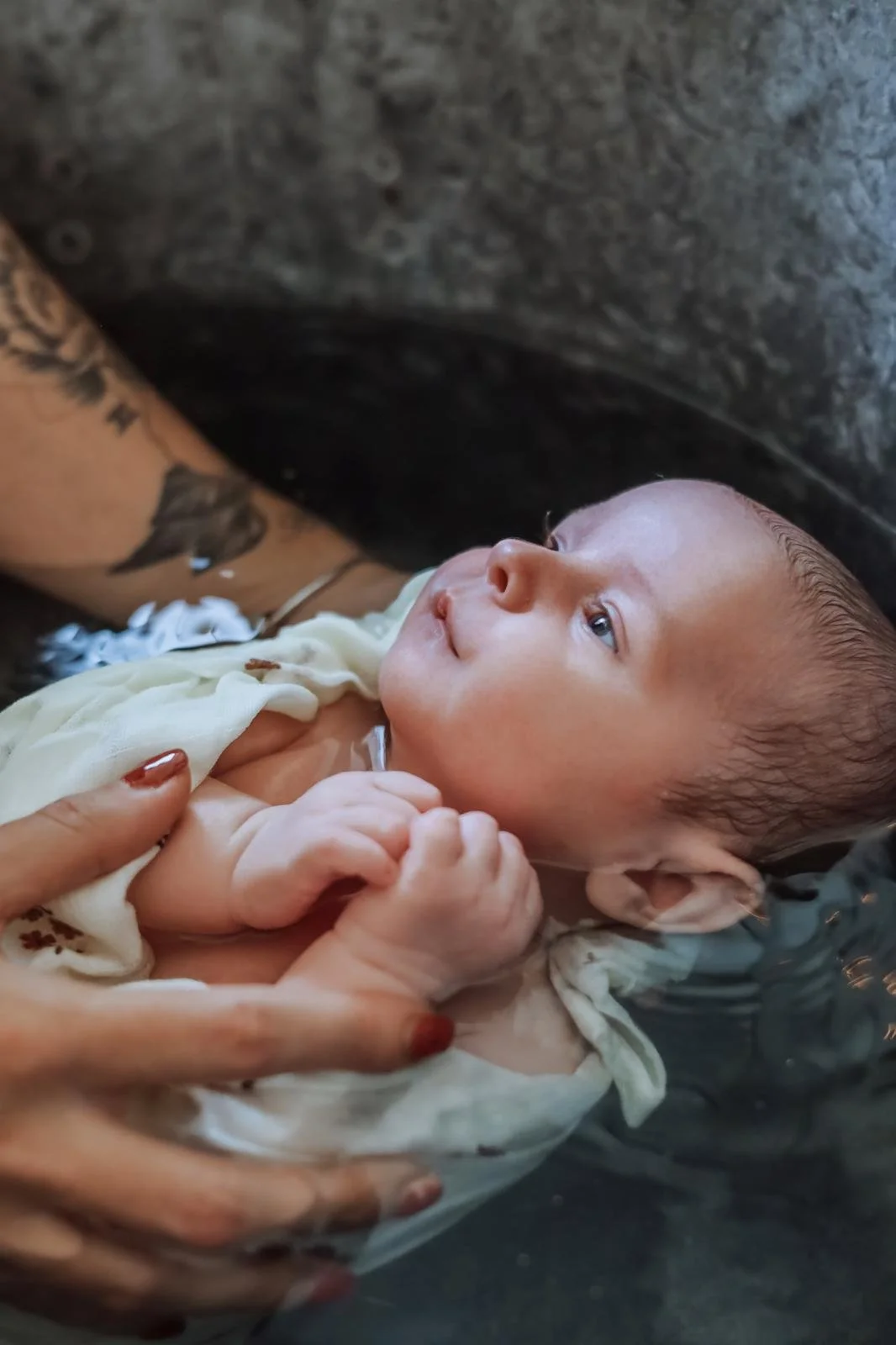 Bébé nageant dans l'eau, soutenu par une personne, avec des tatouages visibles sur le bras. Le bébé regarde vers le haut, l'air calme et curieux.