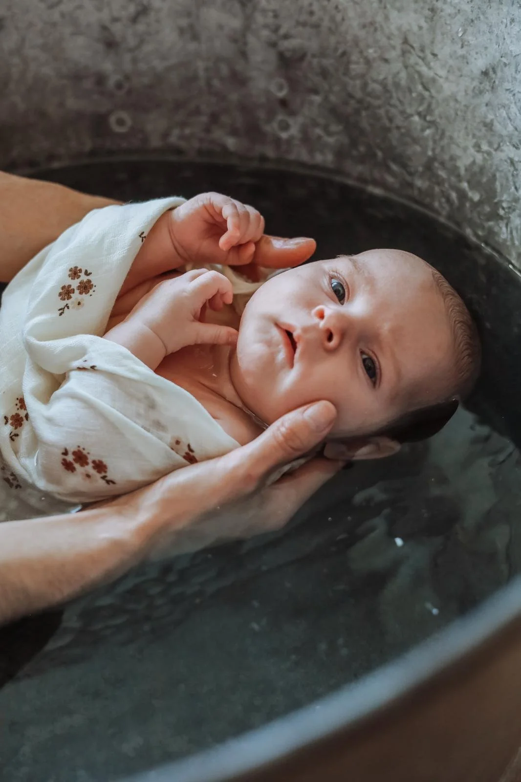 Un bébé est en train d'être baptisé ou de bénéficier d'une bénédiction dans une bassine en métal. La personne tenant le bébé l'immerge doucement dans l'eau, en tenant sa tête et en laissant son visage visible, avec une expression calme et curieuse.