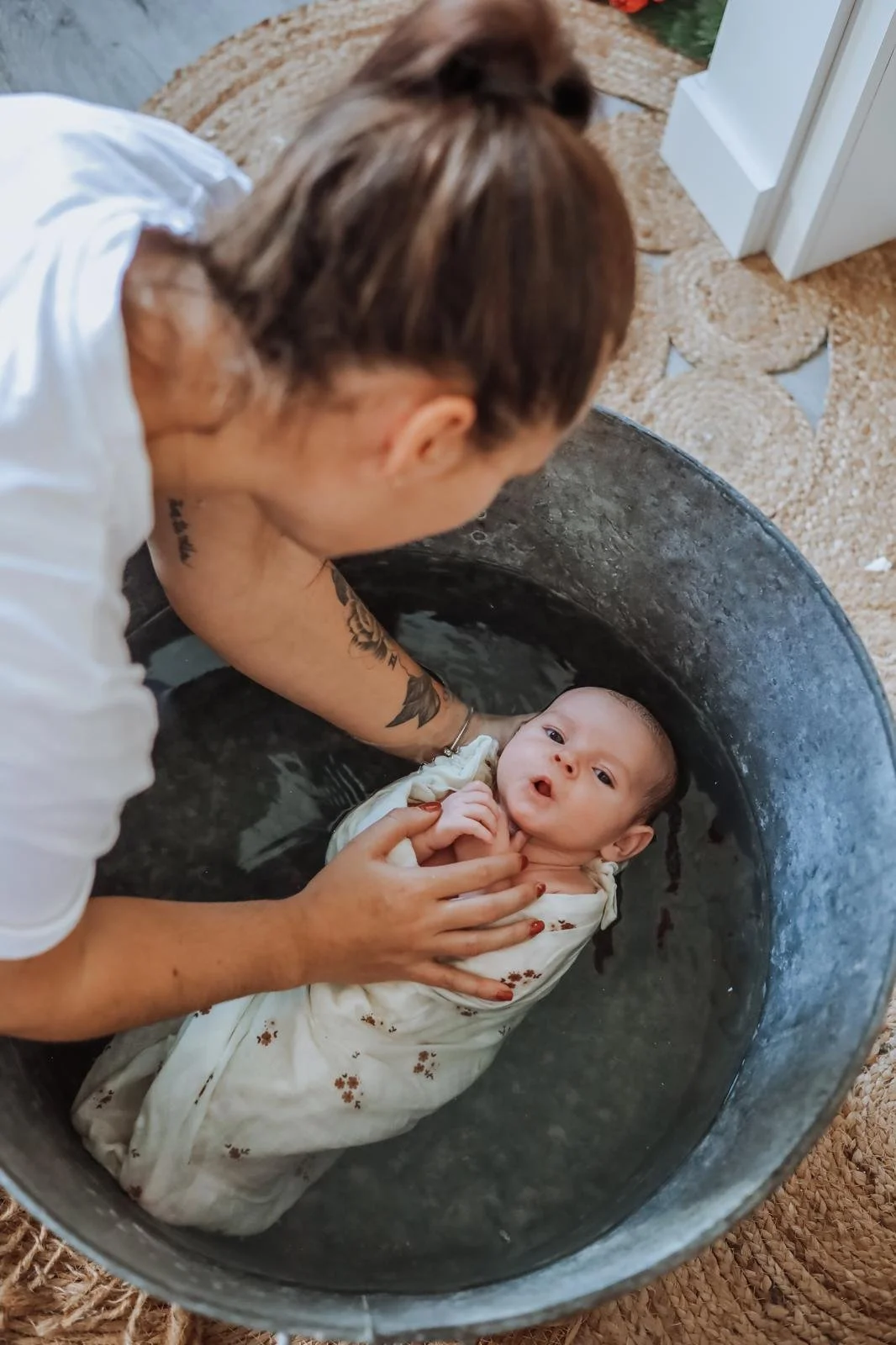 Une femme aide un bébé lors de son baptême dans une grande cuve d'eau, au sol.