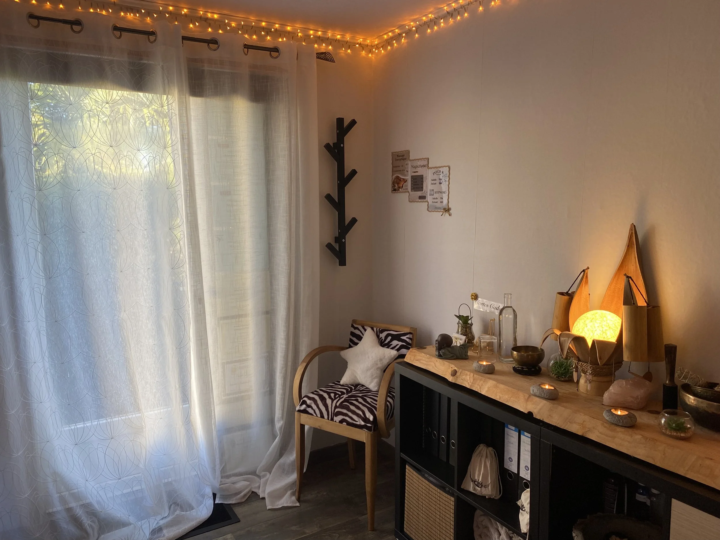 Une pièce cosy, chaleureuse avec un fauteuil en bois, des décorations lumineuses, une étagère murale noire, et une table en bois avec des bougies, des plantes, et une lampe à lumière chaude.