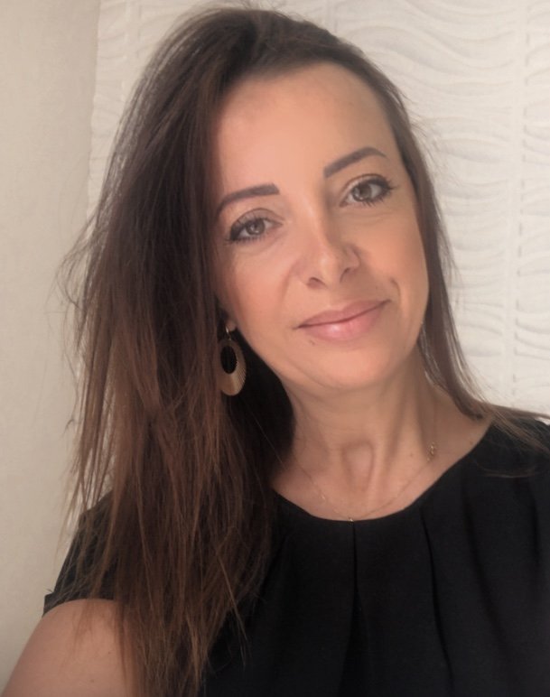 Portrait d'une femme thérapeute, souriante avec des cheveux bruns, portant une robe noire et une boucle d'oreille dorée, devant un mur blanc