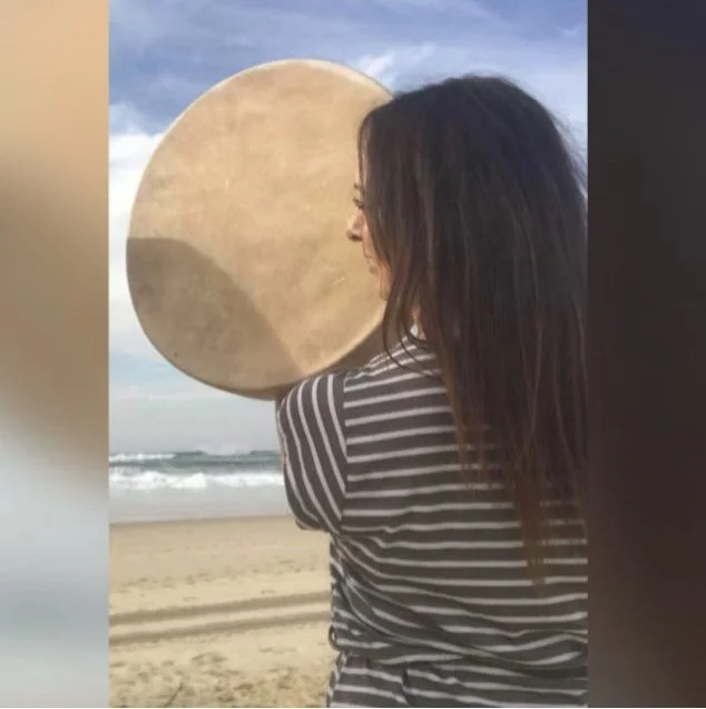 Femme médecine regardant l'océan avec un tambour chamanique en peau de bison devant son visage, créant une illusion d'un corps autour de la tête, sur la plage.