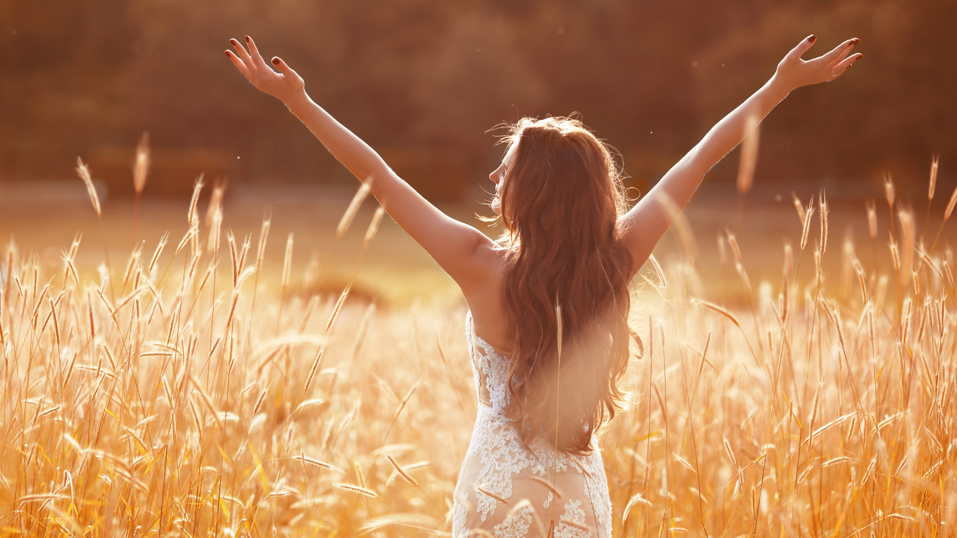 Jeune femme avec des cheveux longs dans un champ de blé, bras levés, à la lumière du coucher de soleil, symbole de liberté.