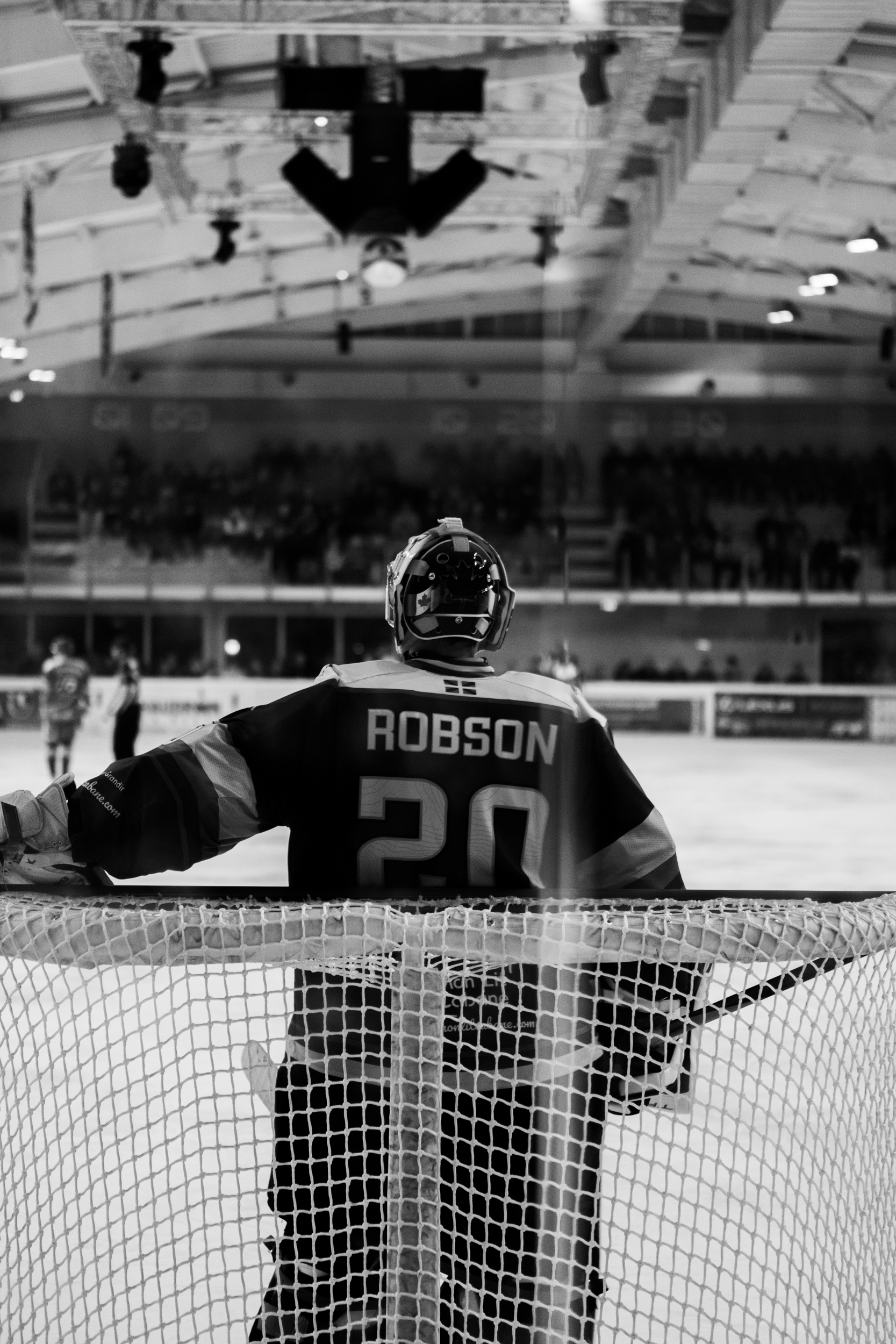 An ice hockey goalie in uniform, number 20, with the name 'Robson' on the back, facing the ice rink during a game, with spectators in the background. Hormadie anglet