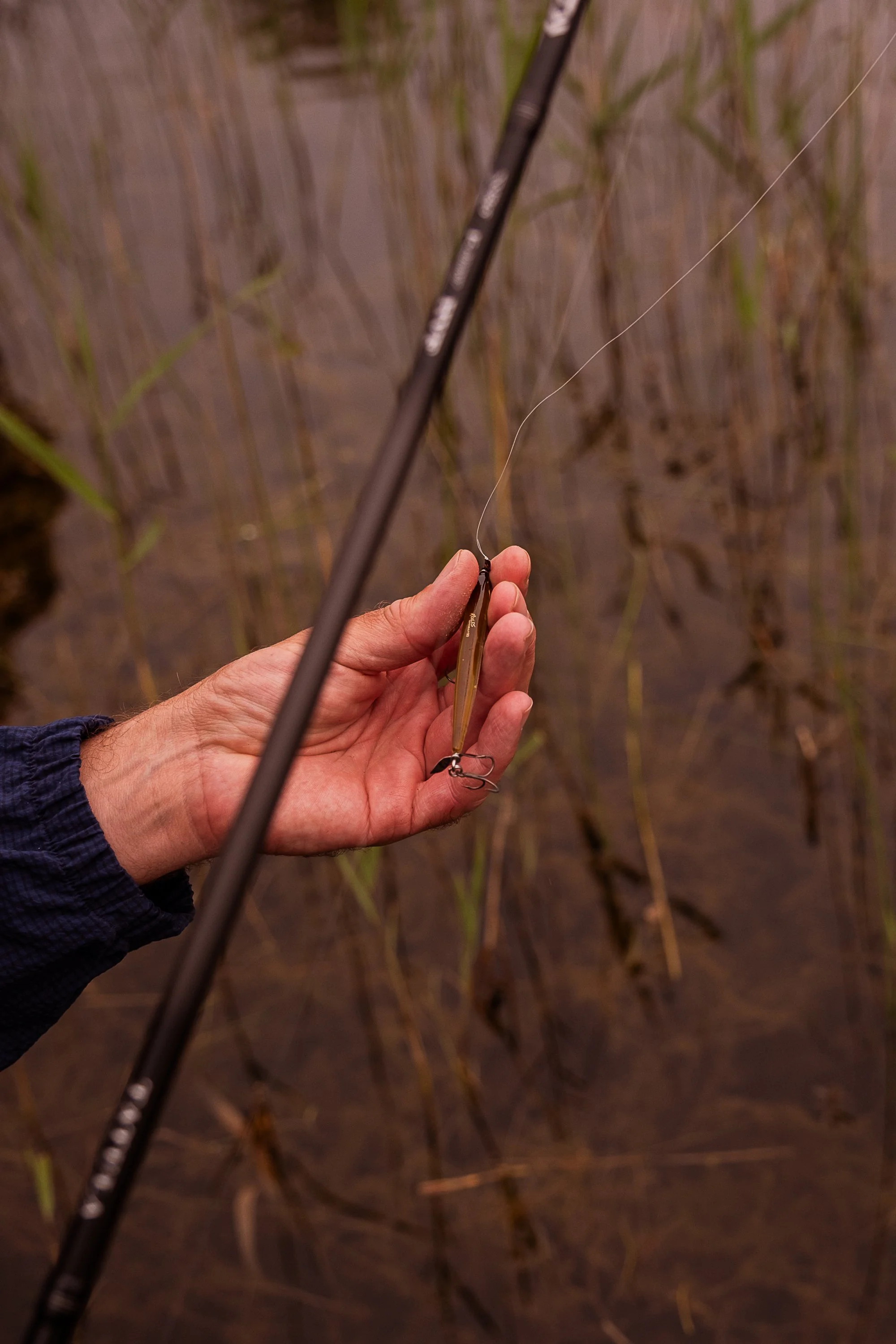 A person holding a fishing lure near water, with a fishing rod visible in the background. River west


