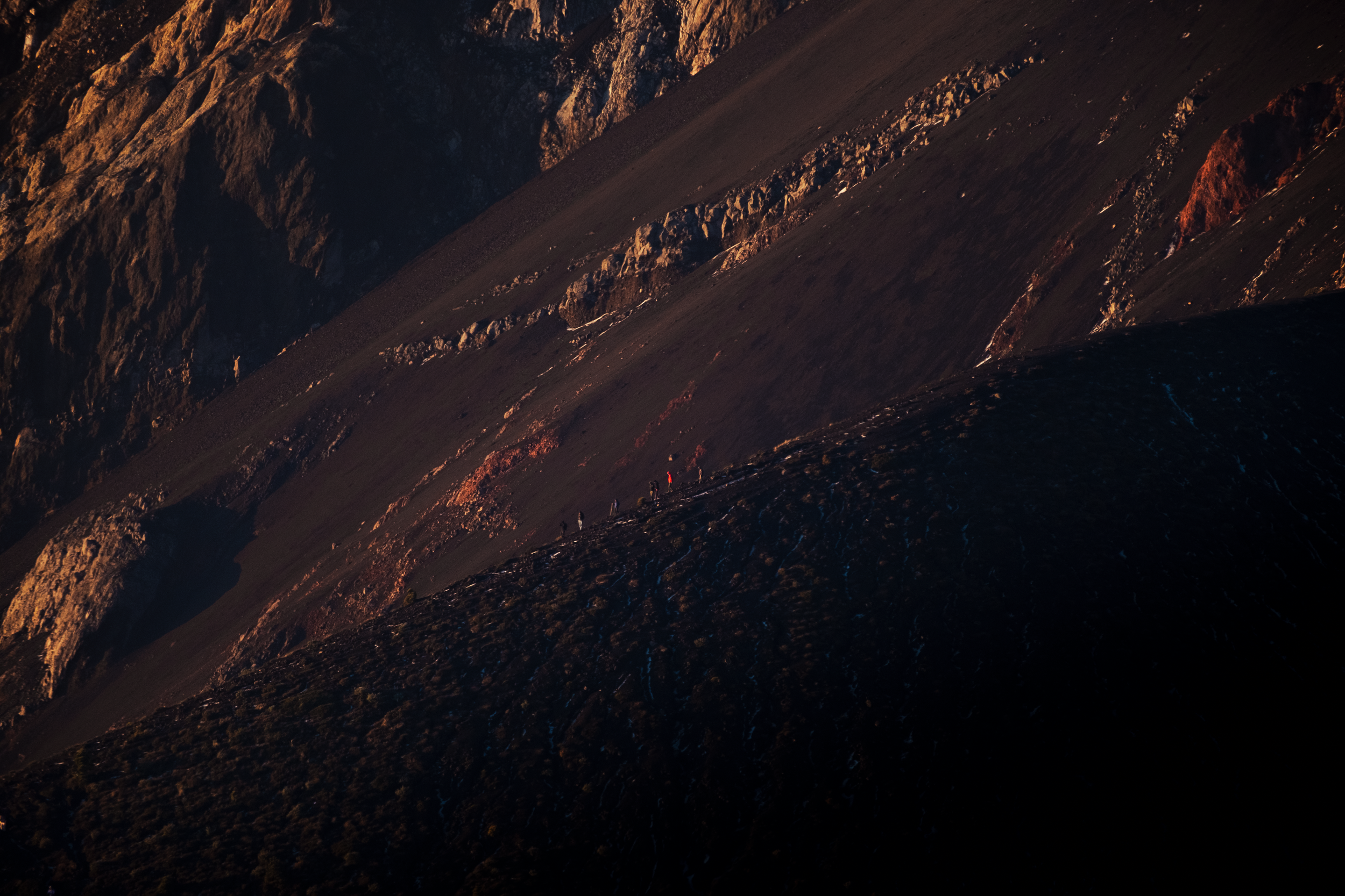 Hikers walking in a line on a mountain ridge during sunrise with steep rocky slopes and shadows on El Fuego volcano
.