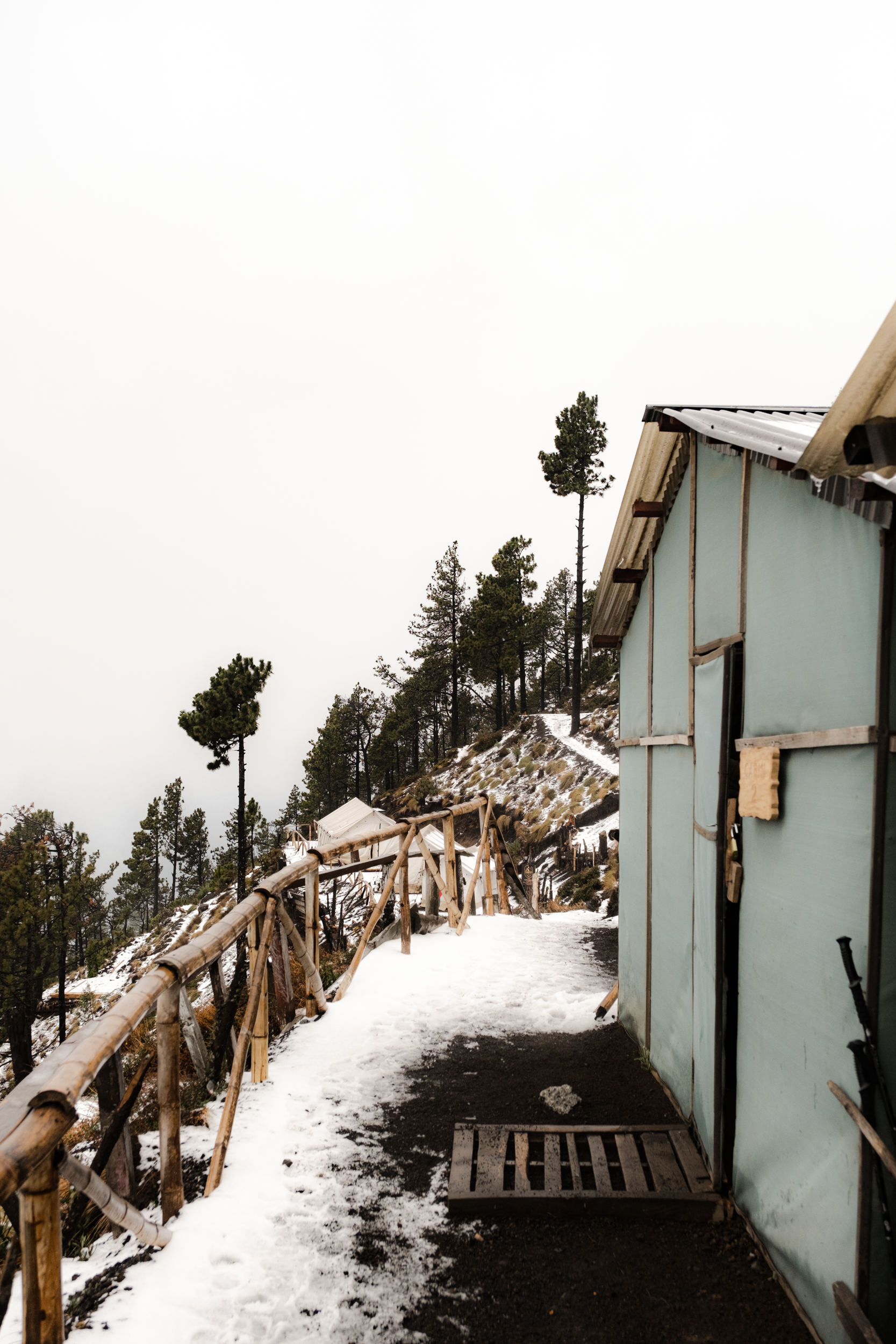 A snowy mountain pathway with a wooden handrail on the left side and a blue building on the right, with tall pine trees in the background. Acatenango, Guatemala