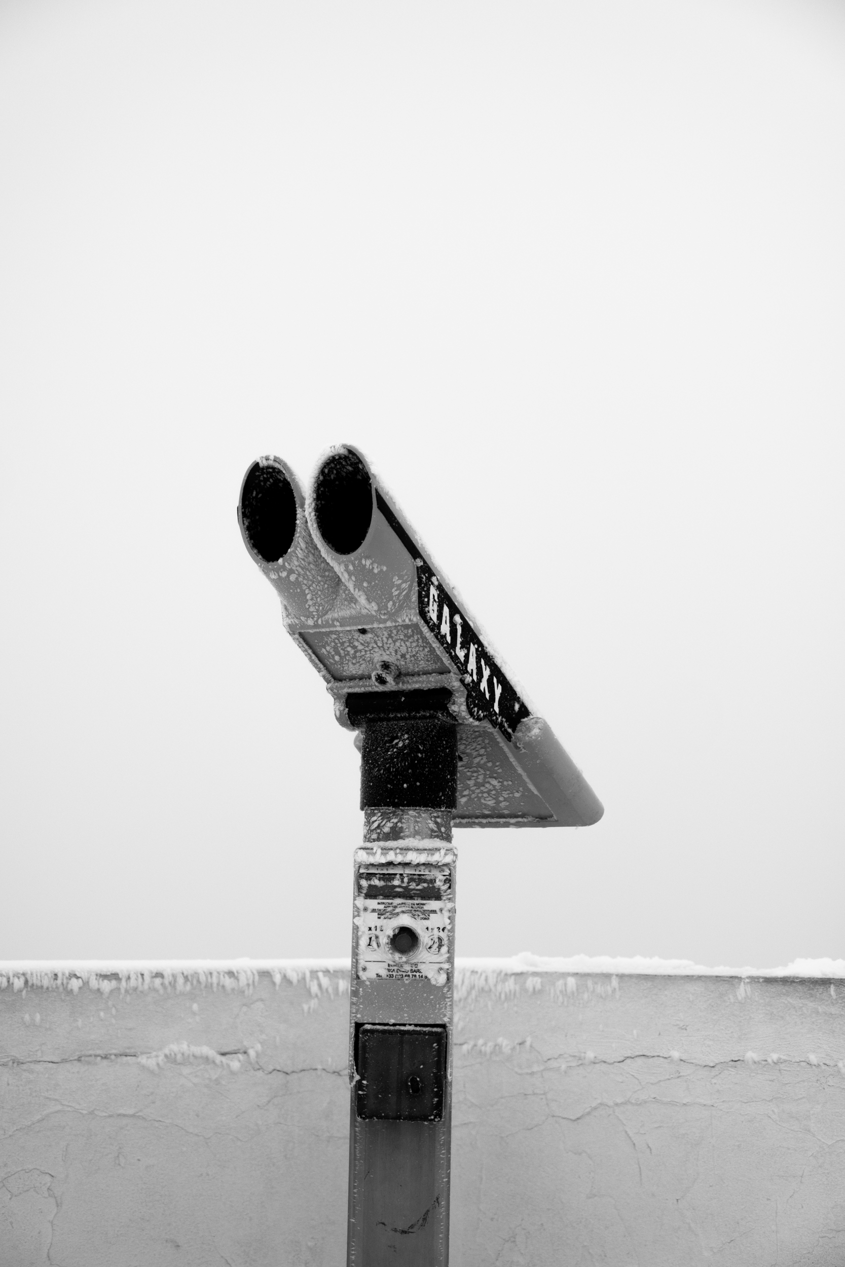 A black-and-white image of a deck of binoculars, mounted on a post, with snow and ice on the surfaces, against a gray sky. La rhune, France