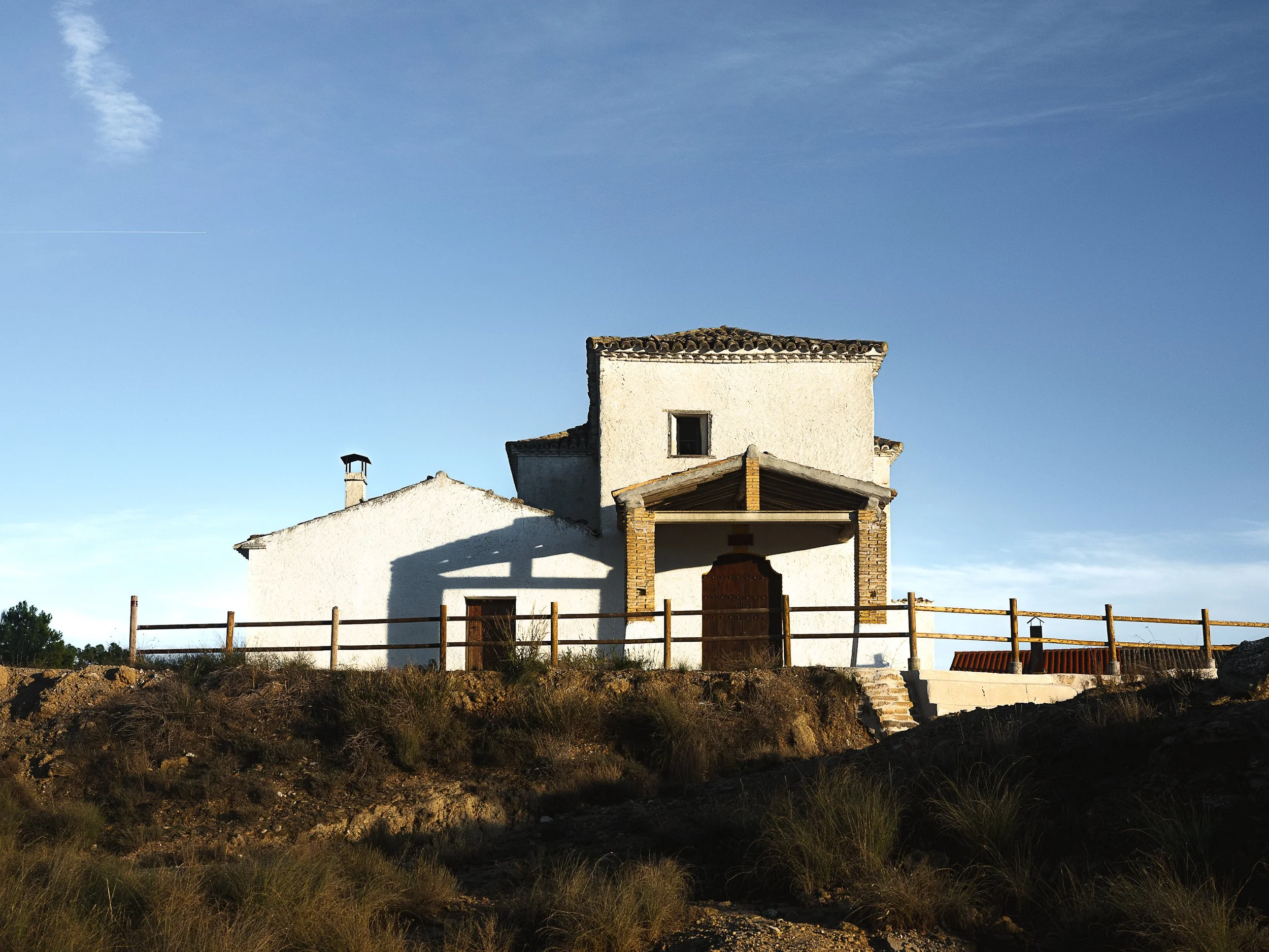 A white house with a sloped tiled roof, situated on a hill with a dirt path leading to its entrance, under a clear blue sky. Monegros desert, Spain 