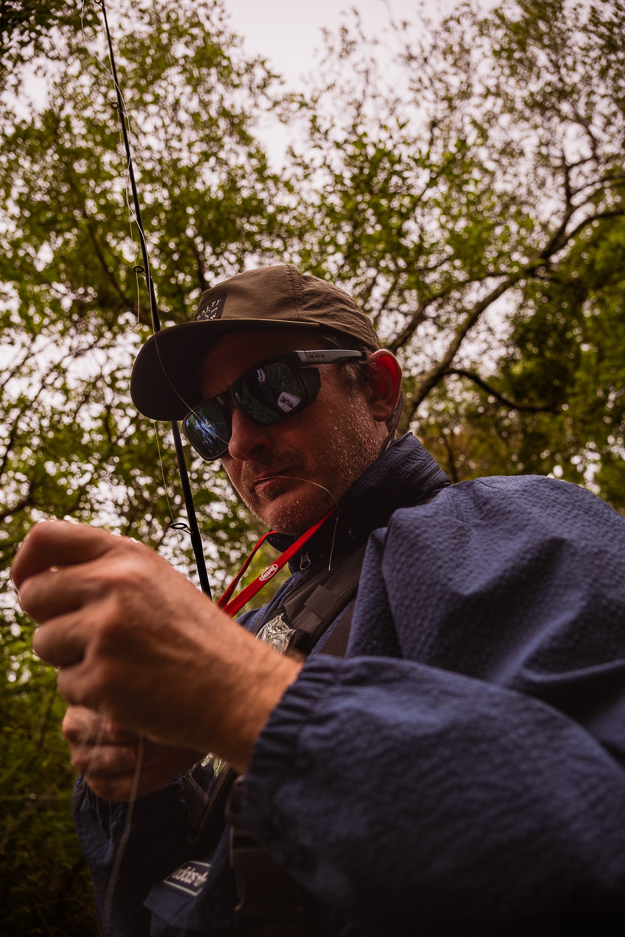 A man wearing a baseball cap, sunglasses, and a dark blue jacket is outdoors, holding a fishing line or string, with trees and a cloudy sky in the background. Work for River West