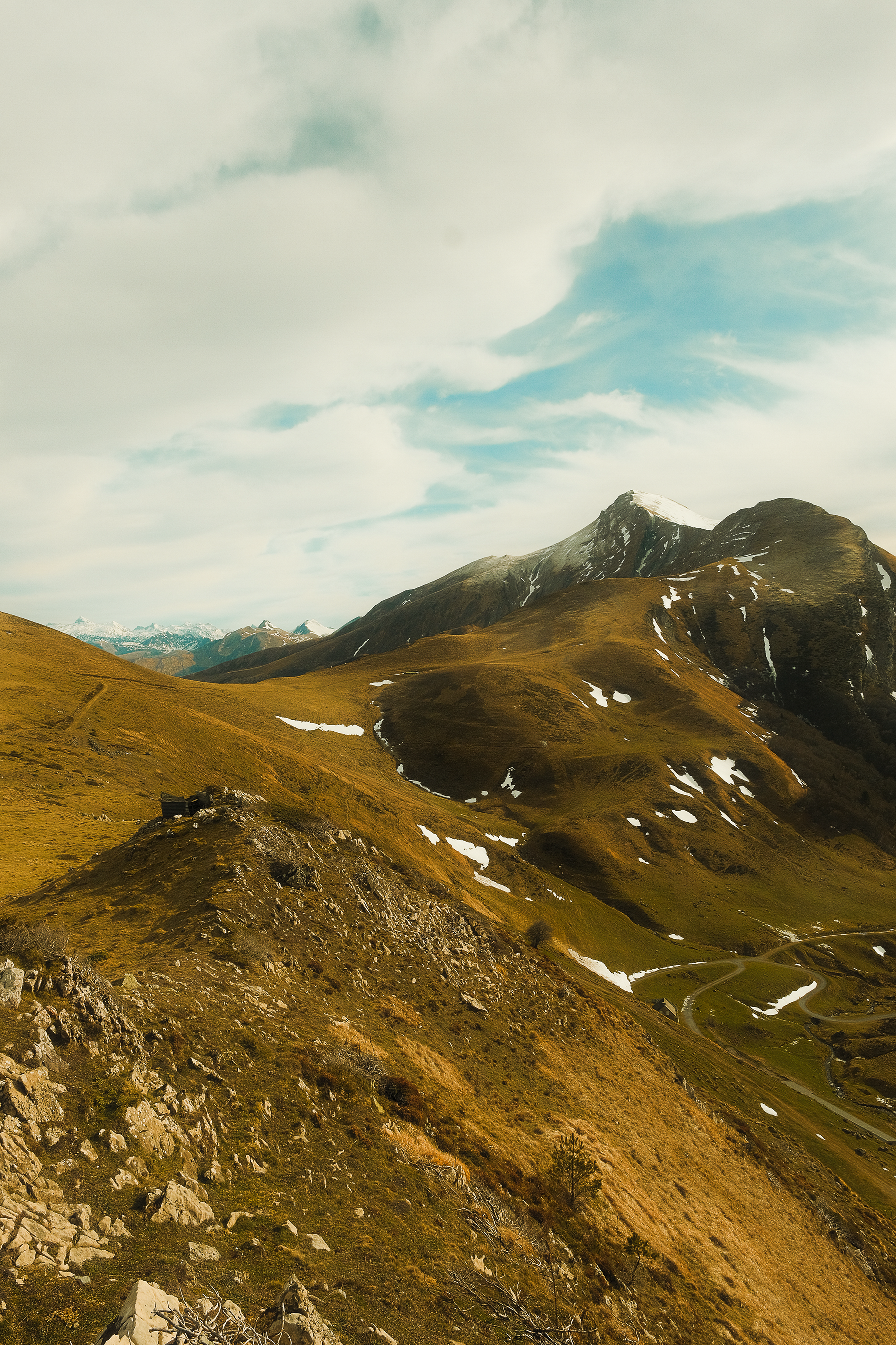 Mountain landscape with grassy slopes, patches of snow, rocky terrain, winding dirt paths, and a partly cloudy sky. Iraty, taken with Fujifilm x-t4
