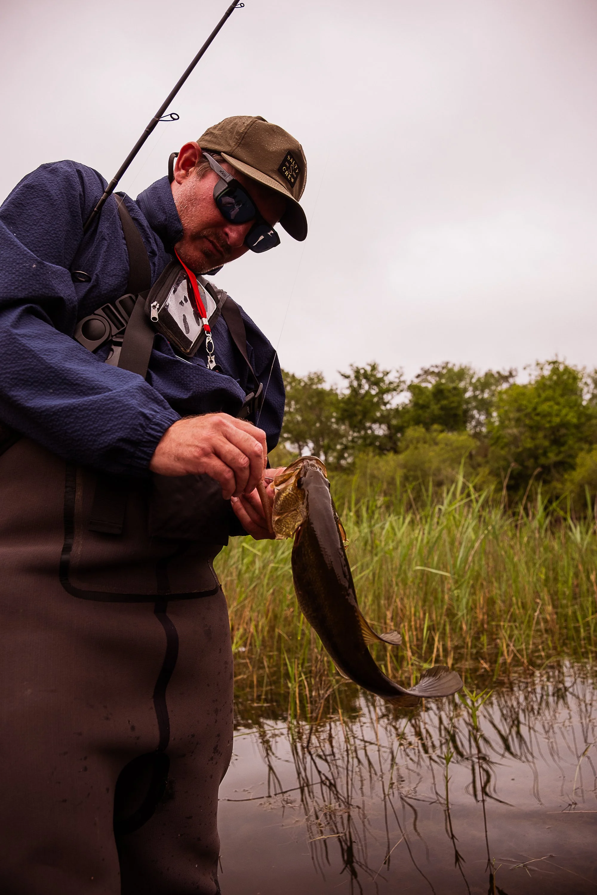 A man wearing sunglasses, a cap, and outdoor gear holding a fish he caught while standing at the edge of a wetland with tall grasses and trees in the background. River West