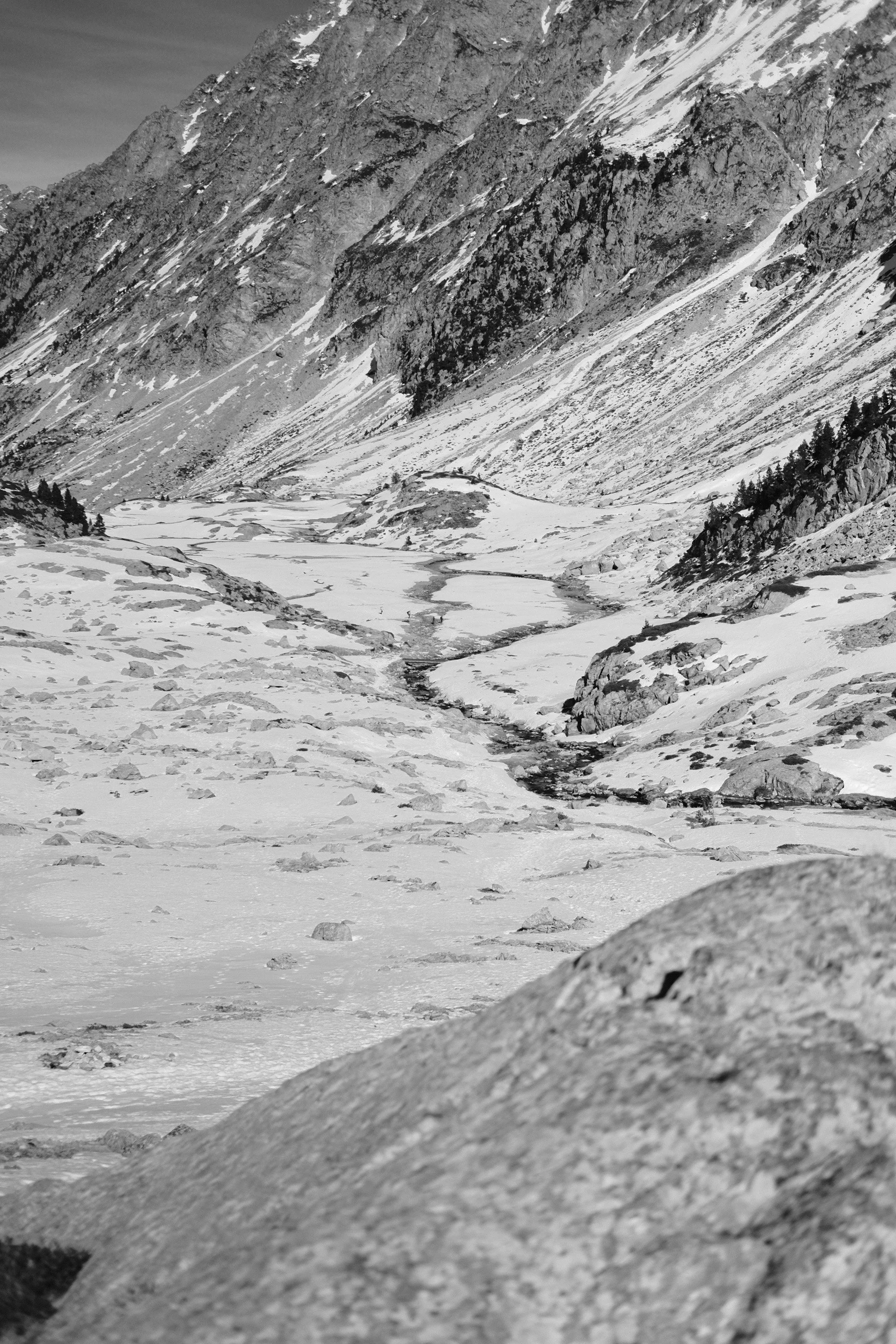 Snow-covered mountain valley with a partially frozen river winding through it, conifer trees on slopes, and mountain peaks in the background. Urth, infra red filter
