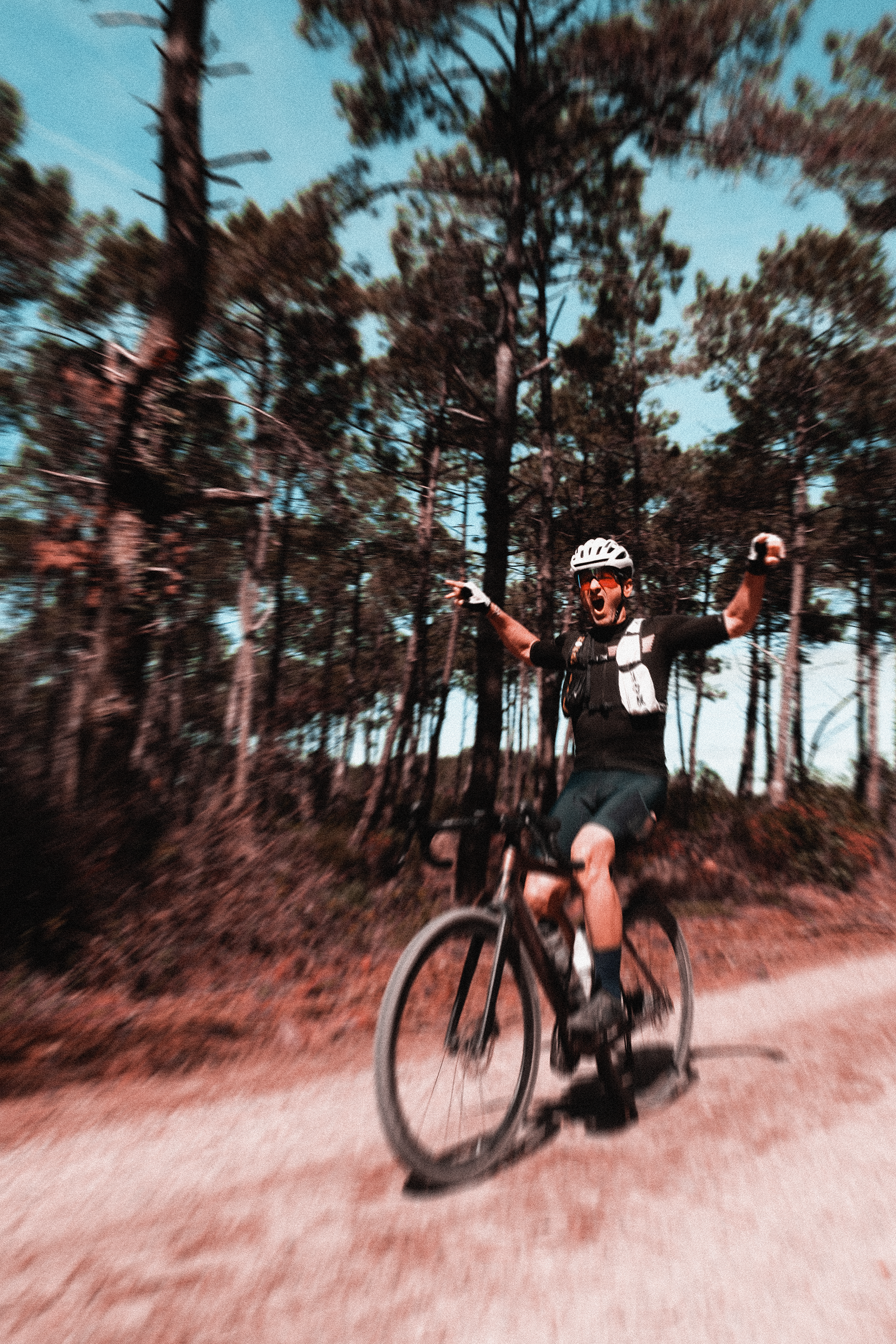 Man riding a mountain bike on a dirt trail through a forest, wearing a helmet and sunglasses, with arms raised in celebration.