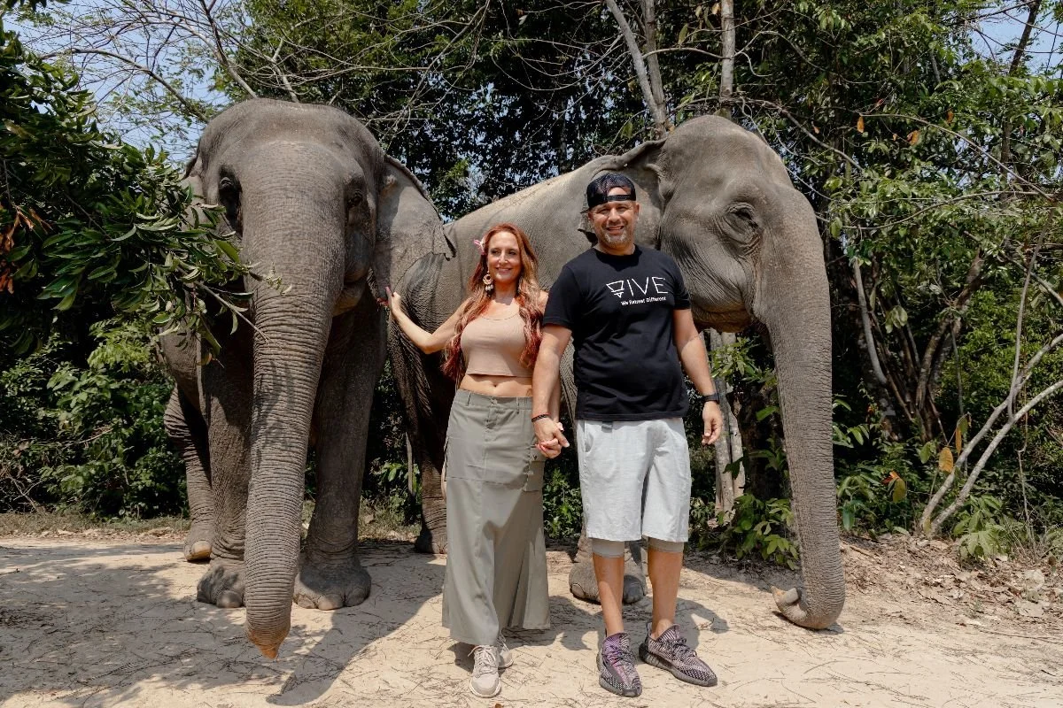 Michelle Ruiz & Nelson Ruiz of VIVE Society Travel visiting an elephant sanctuary in Southeast Asia. Nelson wears a black VIVE logo tshirt and Michelle wears a trendy safari style maxi skirt and tank top.