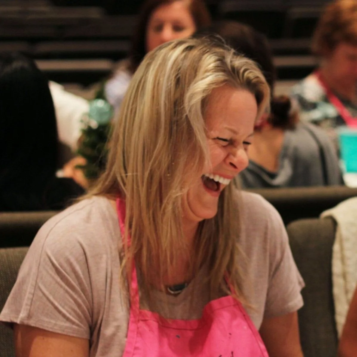 A woman with blonde hair laughing or smiling joyfully at an indoor event, wearing a pink apron.