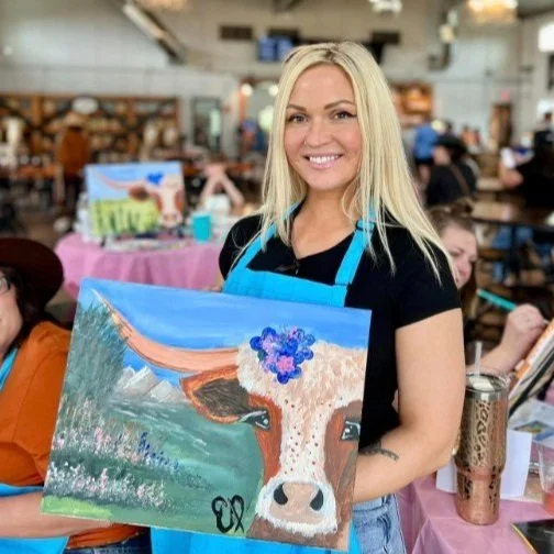 A smiling woman with long blonde hair wearing a black shirt and blue apron, holding a canvas painting of a cow with a scenic background in an art class or workshop.