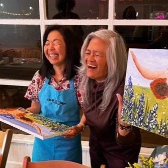Two women standing in a room, smiling and laughing, one holding a children's book and the other holding a colorful painting of a landscape with trees and flowers.