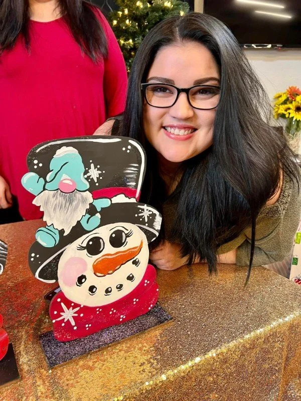 Woman with glasses smiling at a snowman-shaped decorated cookie or cake on a table, with Christmas decorations in the background.