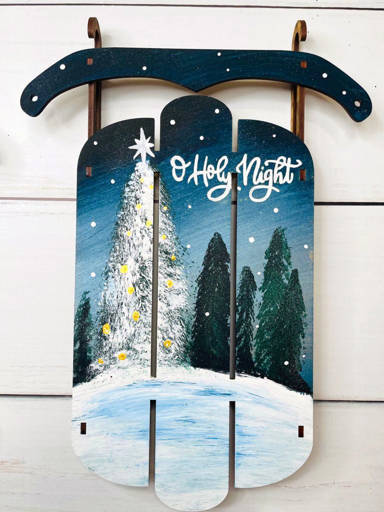 A decorative wooden sign depicting a winter scene with a snow-covered tree decorated with yellow lights, dark evergreen trees, and a snowy ground under a starry night sky. The sign has the words 'O Holy Night' written on it.