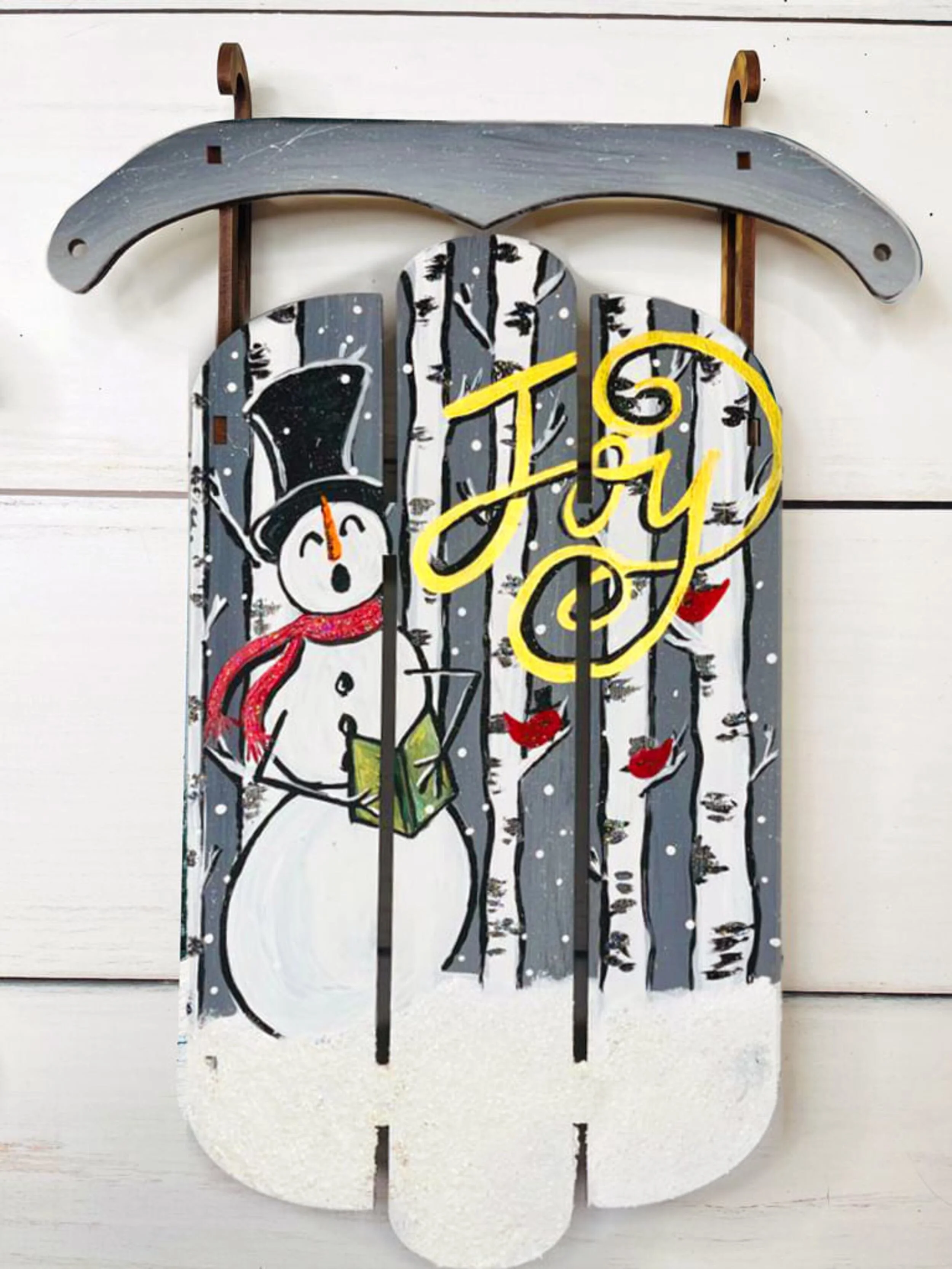 Decorative wall hanging with a snowman wearing a top hat and red scarf, in a winter forest scene with snow and small red birds, and the word "Joy" in yellow script.