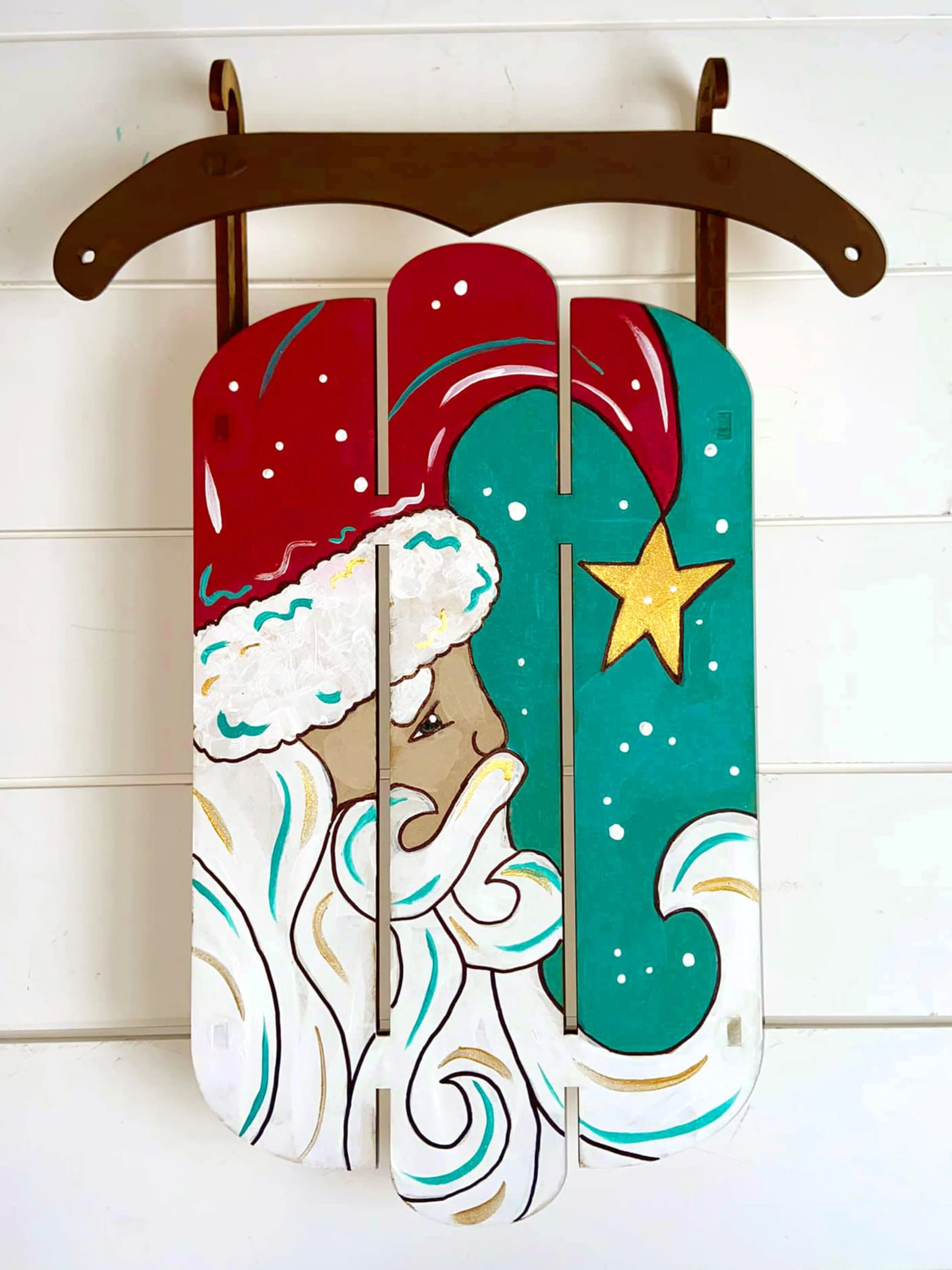 Painted wood decoration featuring Santa Claus in a red hat with white trim and a beard, with a yellow star hanging in the background, on a white wooden wall.