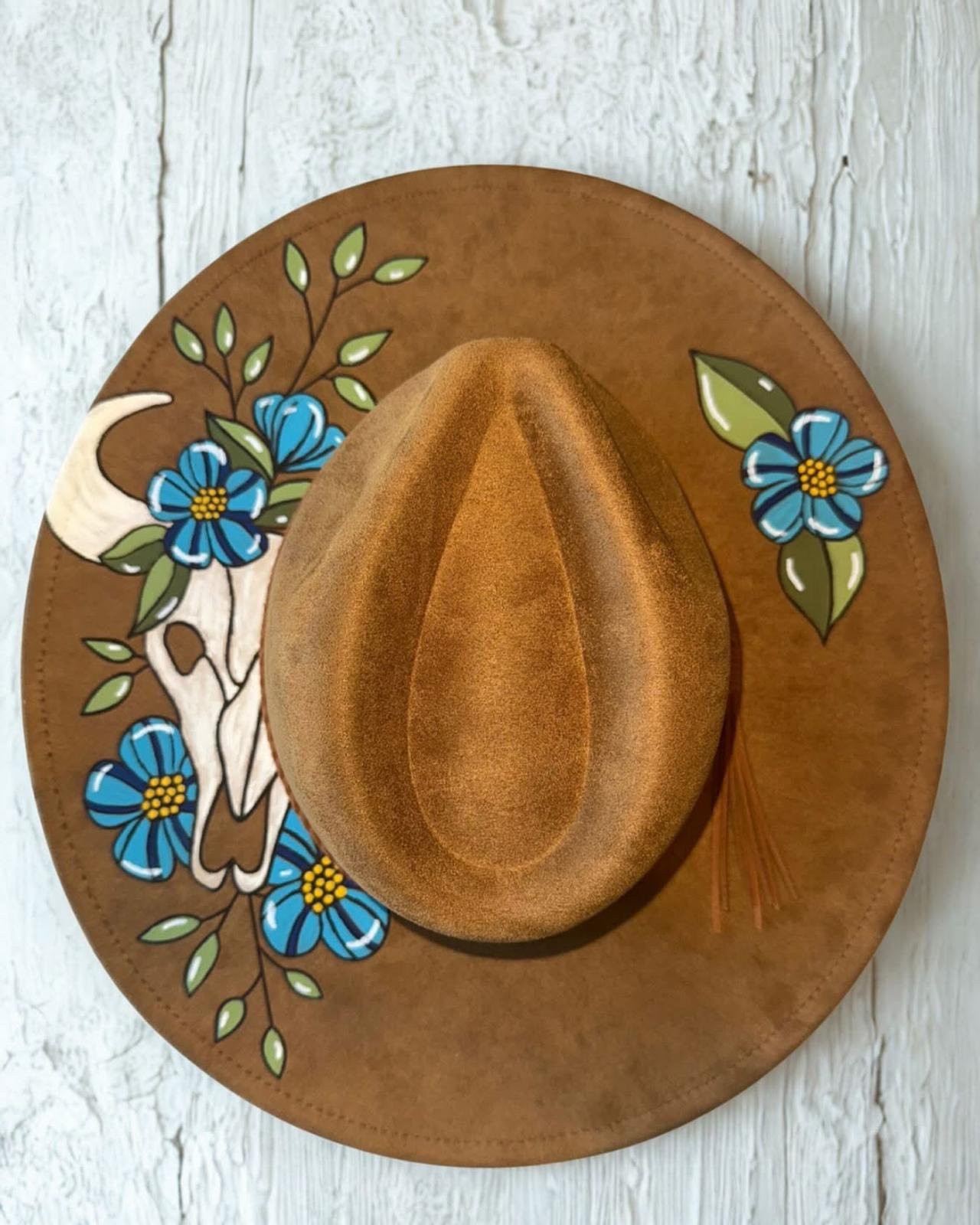 A brown cowboy hat with a decorative band featuring blue flowers and green leaves, placed on a painted round wooden surface with a cow skull illustration.