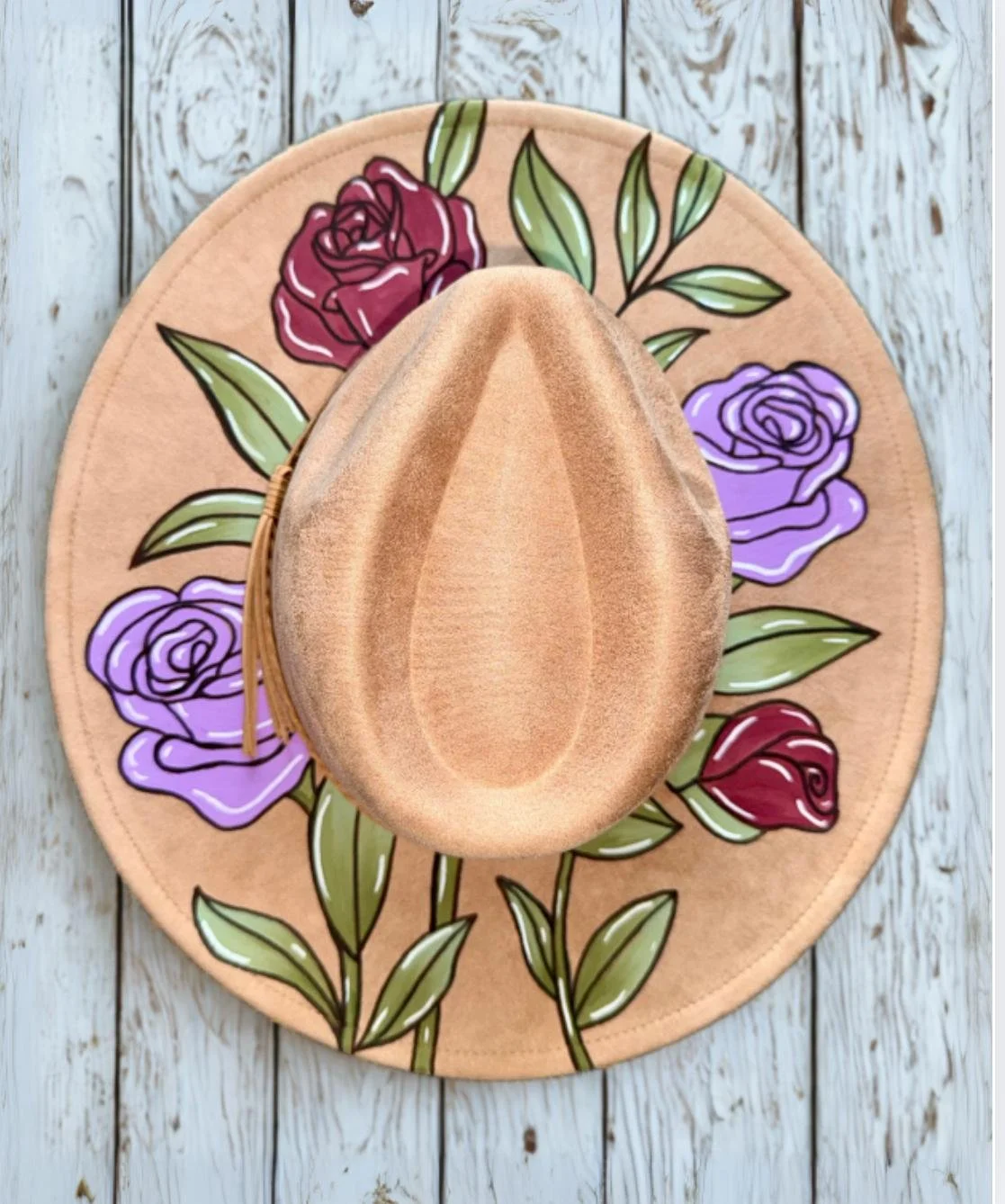 A tan straw hat with a wide brim, placed on a round floral cloth featuring painted roses in red and purple with green leaves, on a white wooden surface.