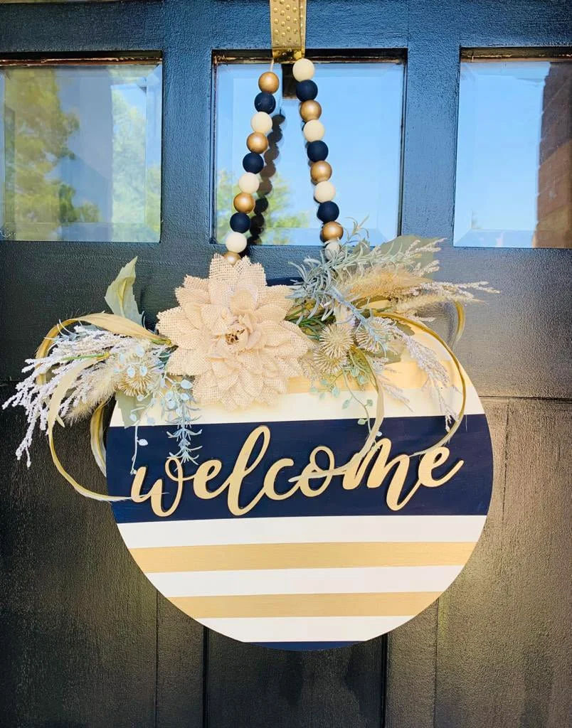 A decorative welcome sign hanging on a black door with window panes, decorated with flowers, foliage, and a string of beads.