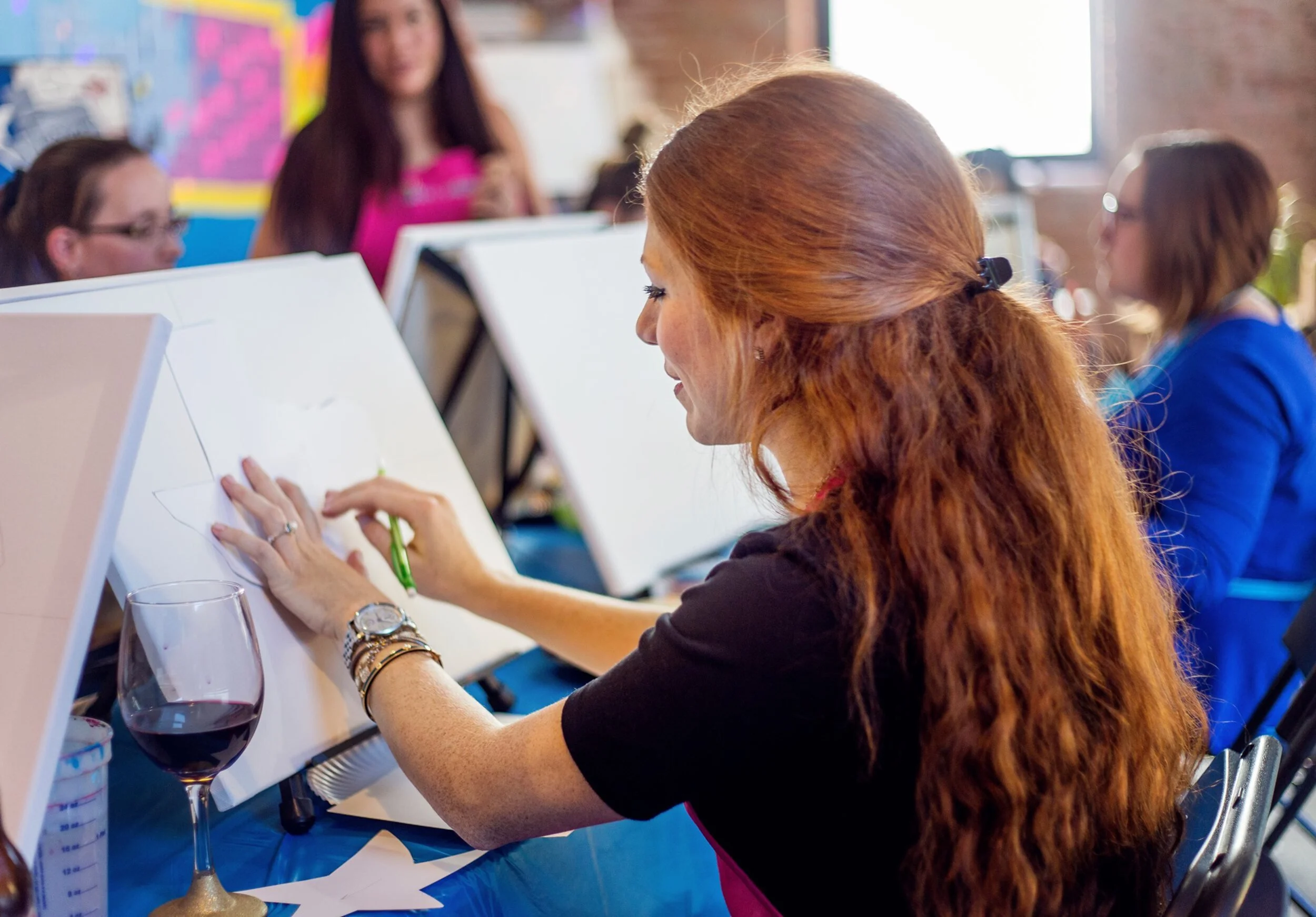 A woman with long red hair drawing a star on a white canvas at an art event, with a glass of red wine on the table, and several other women in the background