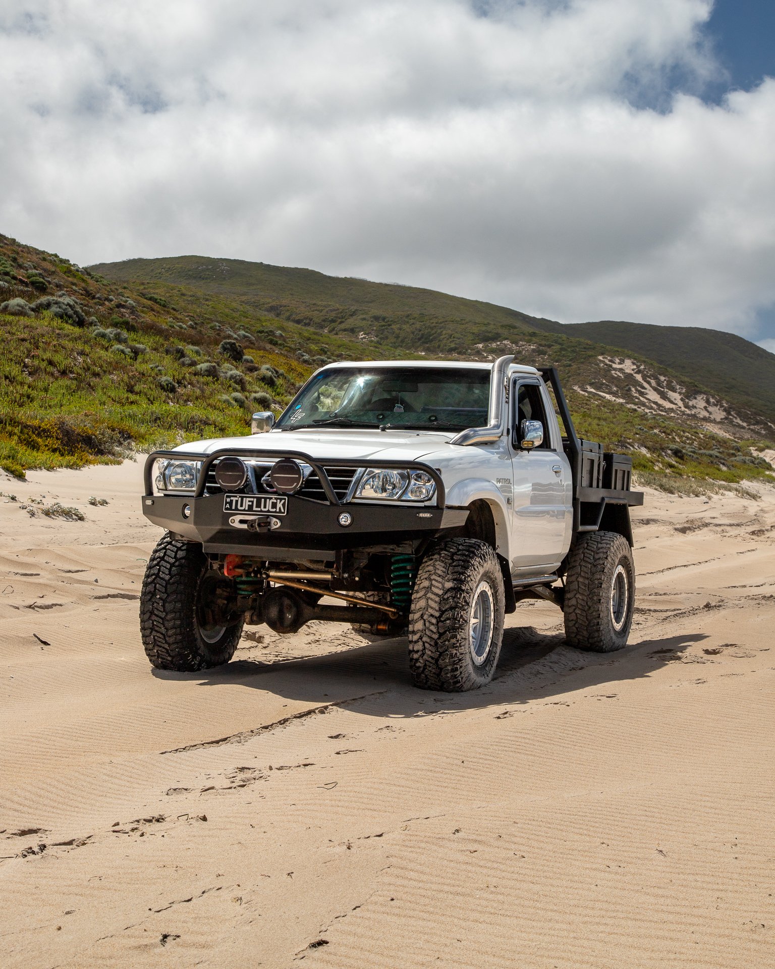 A white GU Patrol ute parked on sandy terrain with green hills and cloudy sky in the background.