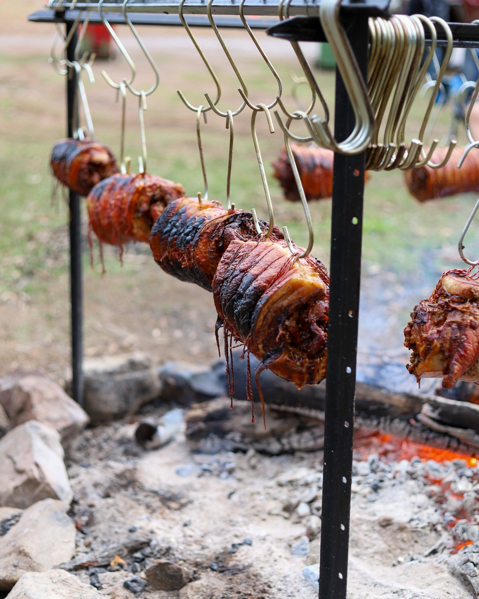 Pork pieces hanging on hooks over a bed of ashes and a small fire for outdoor smoking or roasting.