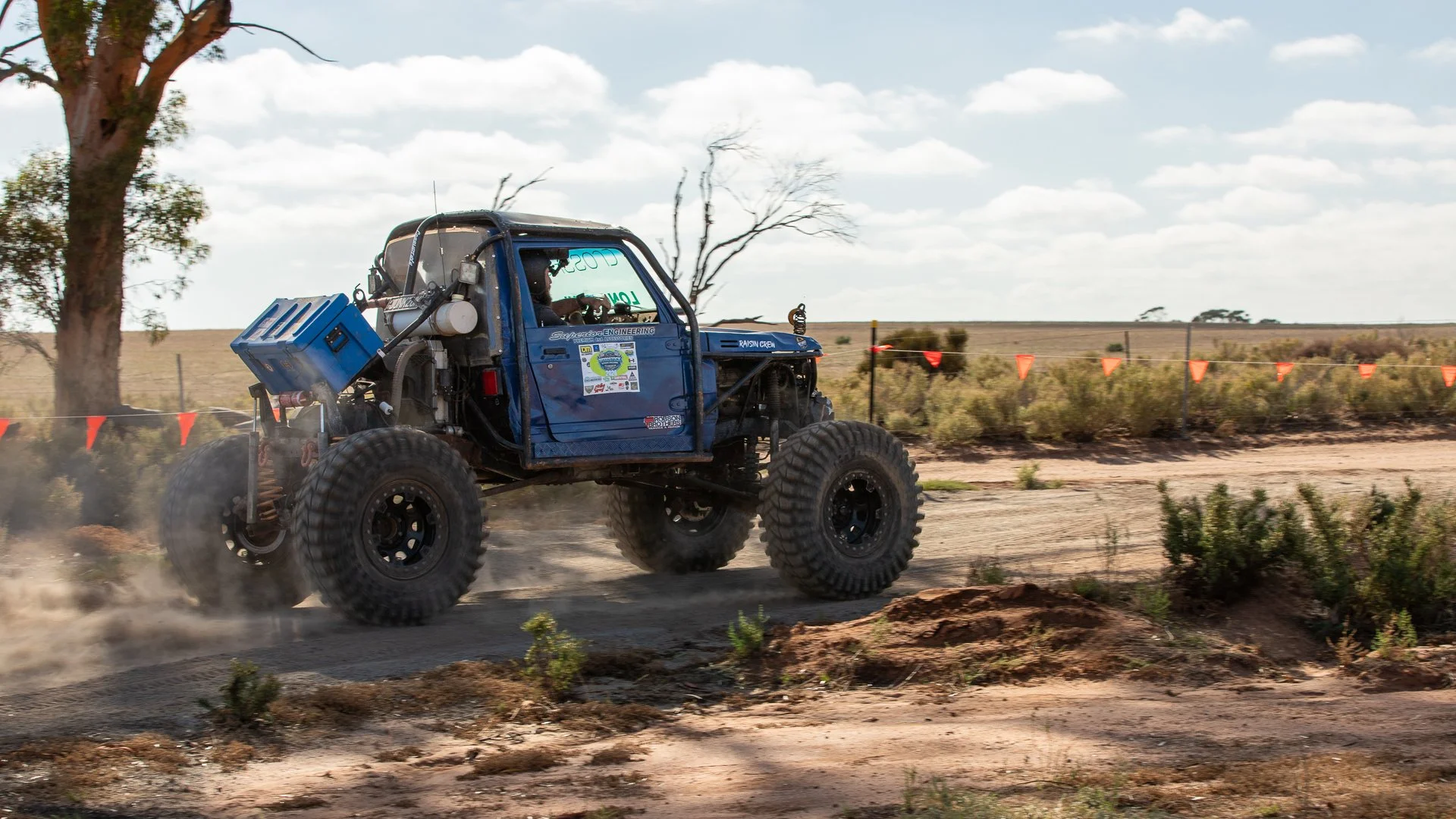A blue 4wd comp truck driving on a dirt track in a desert landscape with sparse vegetation and a clear sky.