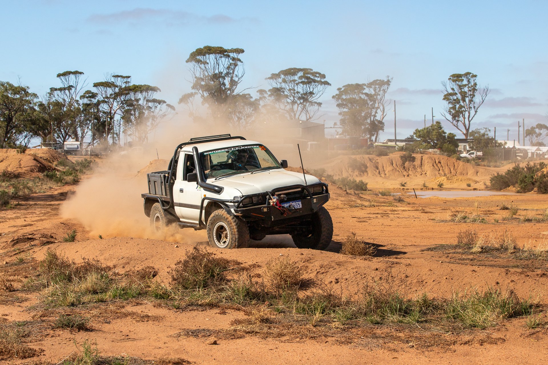 A white 4WD driving on a dusty terrain with dust clouds behind it, surrounded by sparse vegetation and trees in the background.