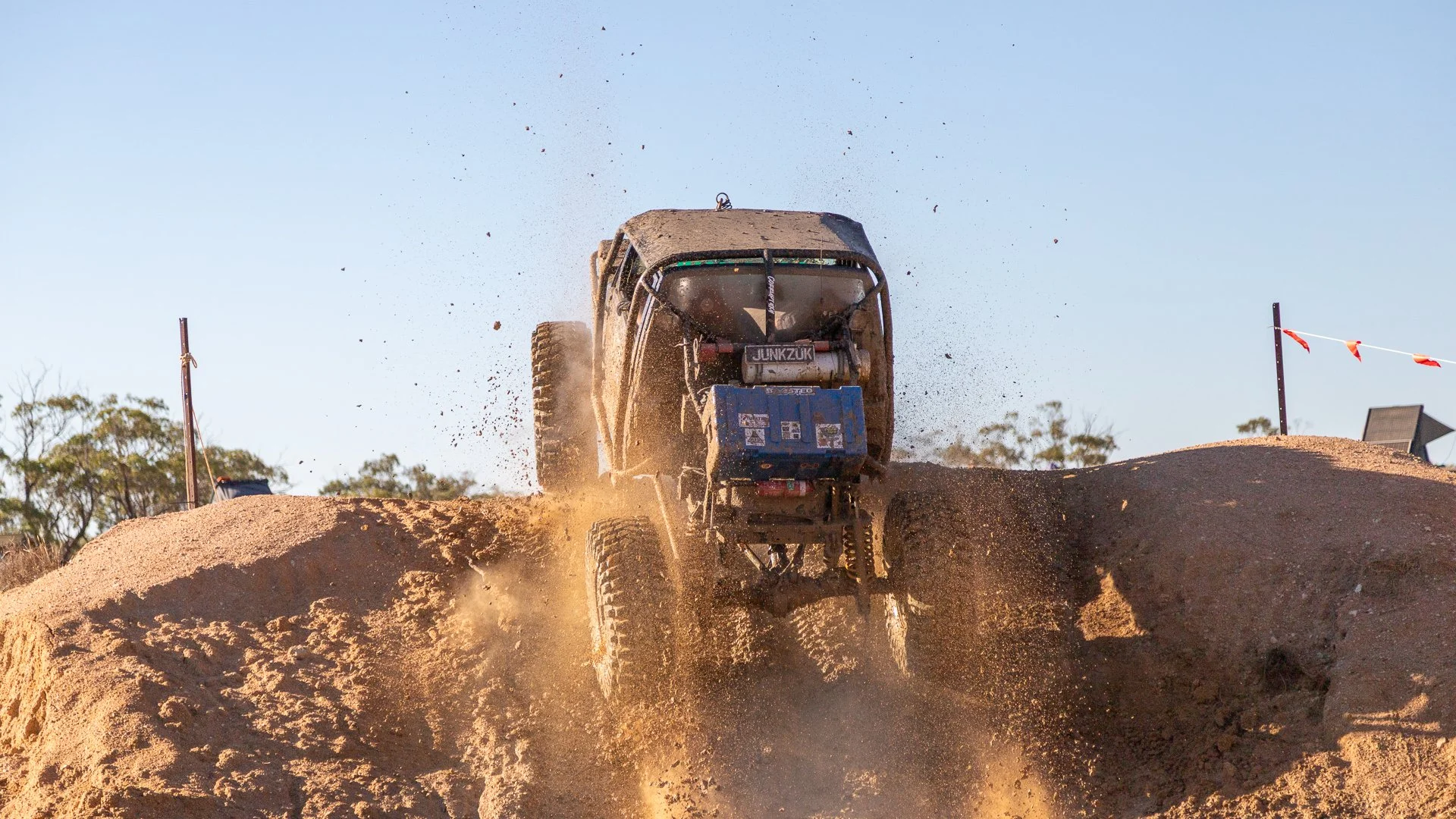 A 4wd comp truck jumping over a dirt mound during a race, kicking up dust with trees and flags in the background during daylight.