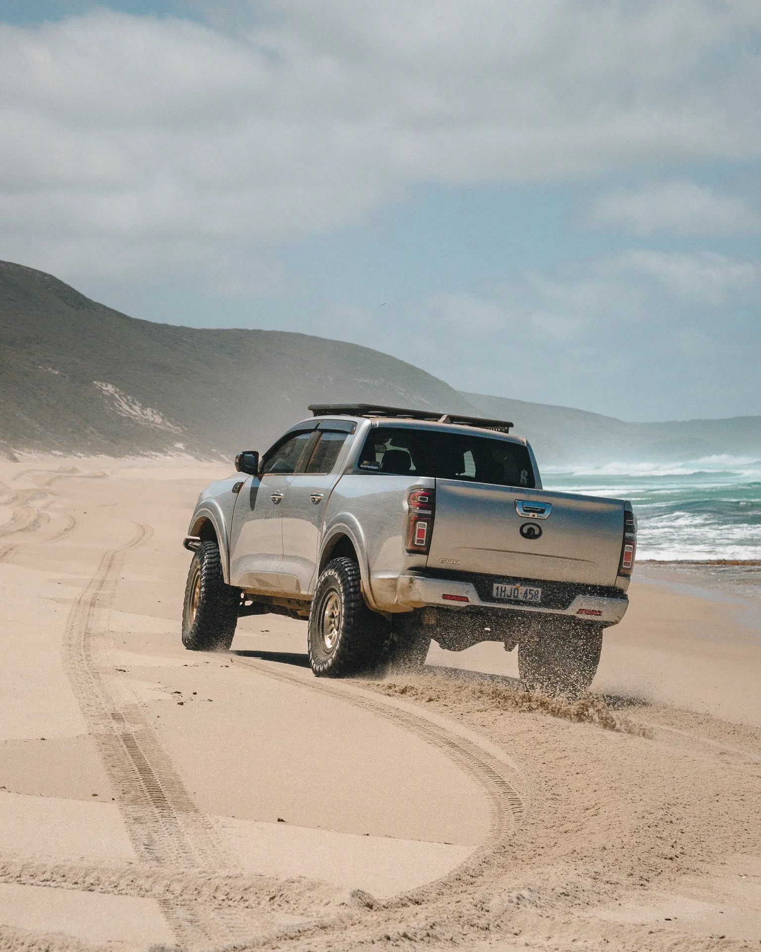 A silver GWM Cannon driving along a sandy beach near the ocean with hills in the background.