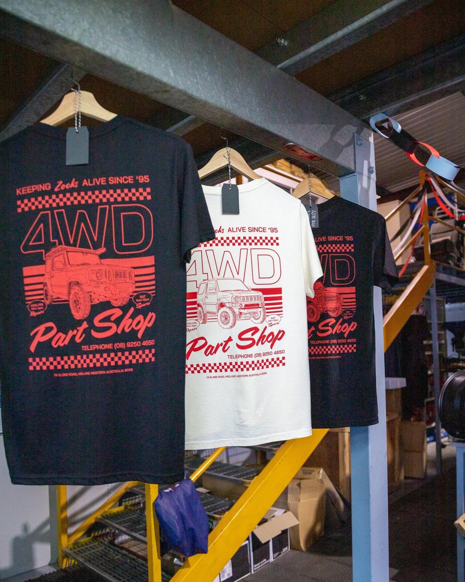 Three 4WD Part Shop T-shirts hanging on a rack, featuring an illustration of different generations of Suzuki Jimnys.
