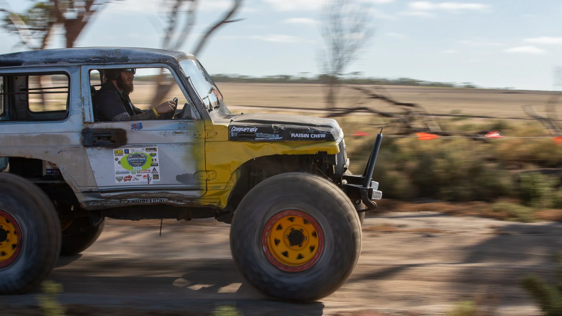 A rugged off-road vehicle with a racing number and decals, driven by a person wearing sunglasses and a helmet, speeding through a desert landscape with blurred background.