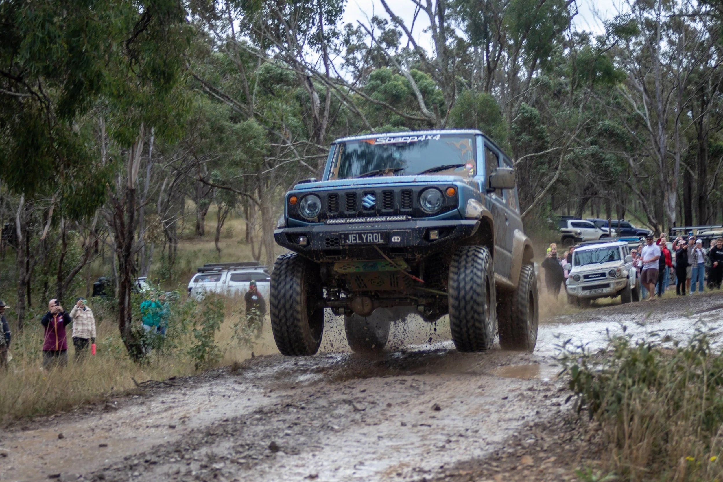 Jelyrol, a blue JB74 Suzuki Jimny jumps over a dirt trail during an offroad speed run event. Spectators stand along the side of the trail watching and taking photos amid a wooded area with tall trees.