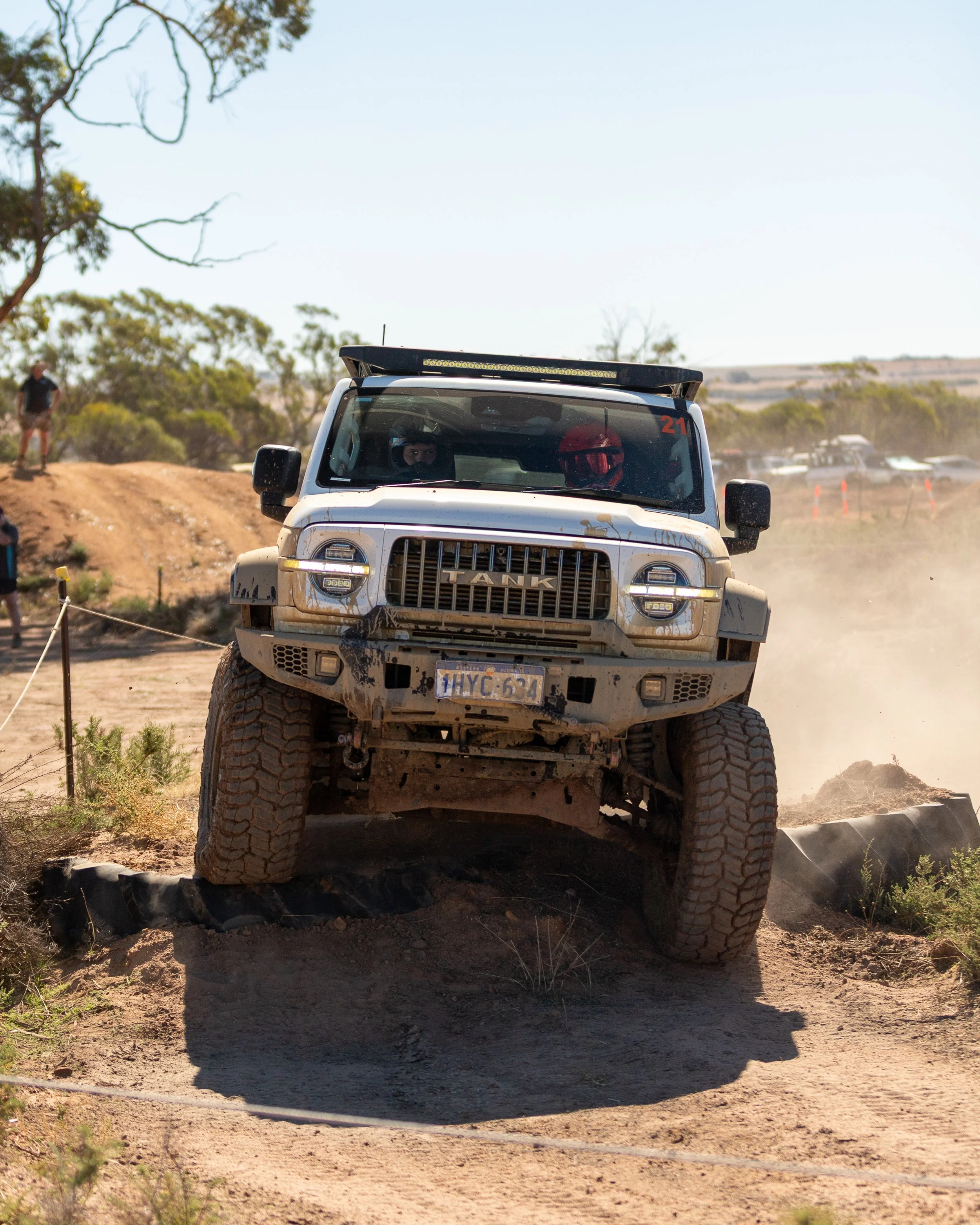 A white off-road vehicle, a Toyota Tundra, navigating a rugged dirt trail in a desert landscape during daytime, with dust kicking up behind it. The vehicle has mud on the front and a roof rack, and there are people and other vehicles in the background.