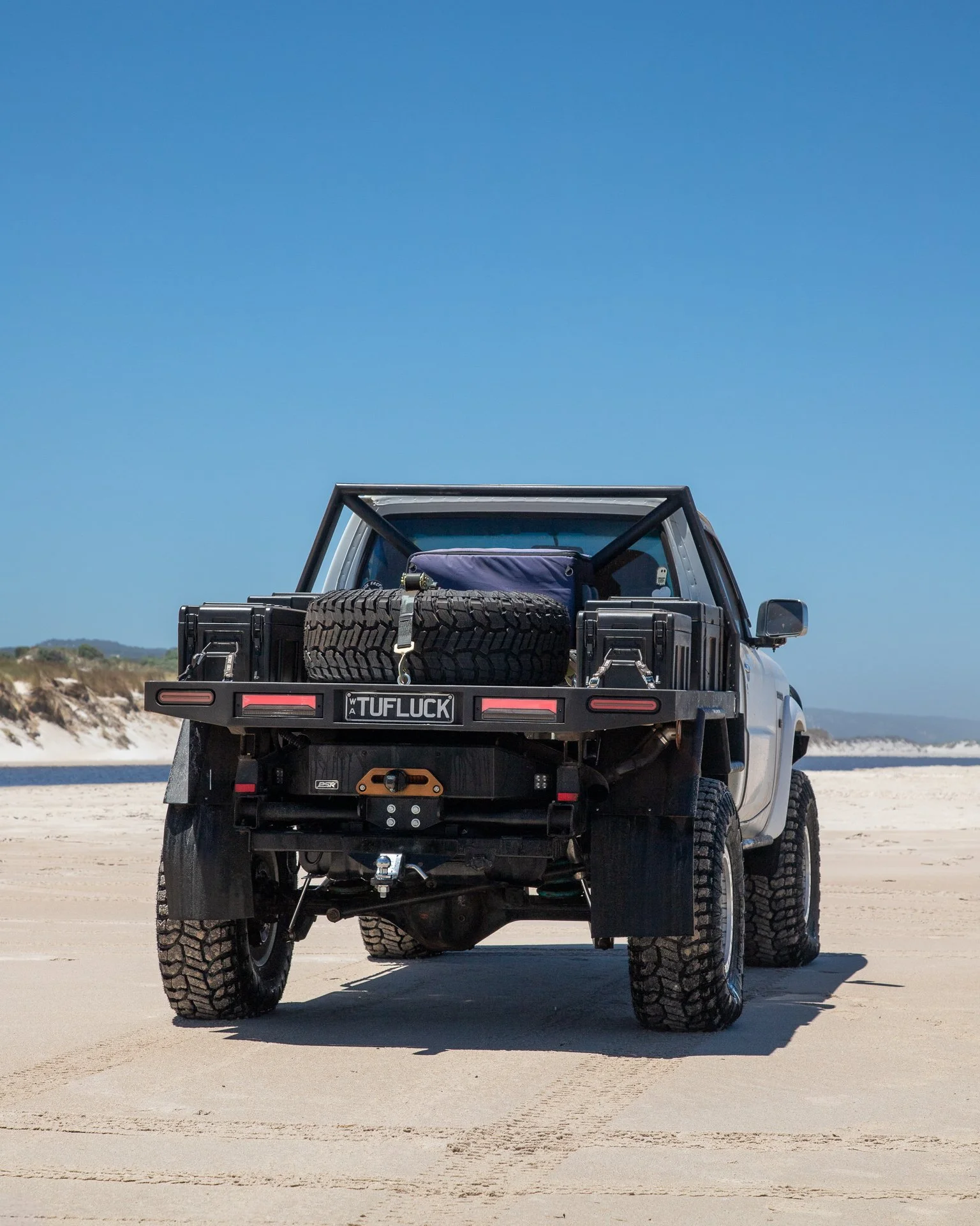 A GU Patrol ute parked on a sandy beach under a clear blue sky, carrying a spare tire and equipment in its bed.