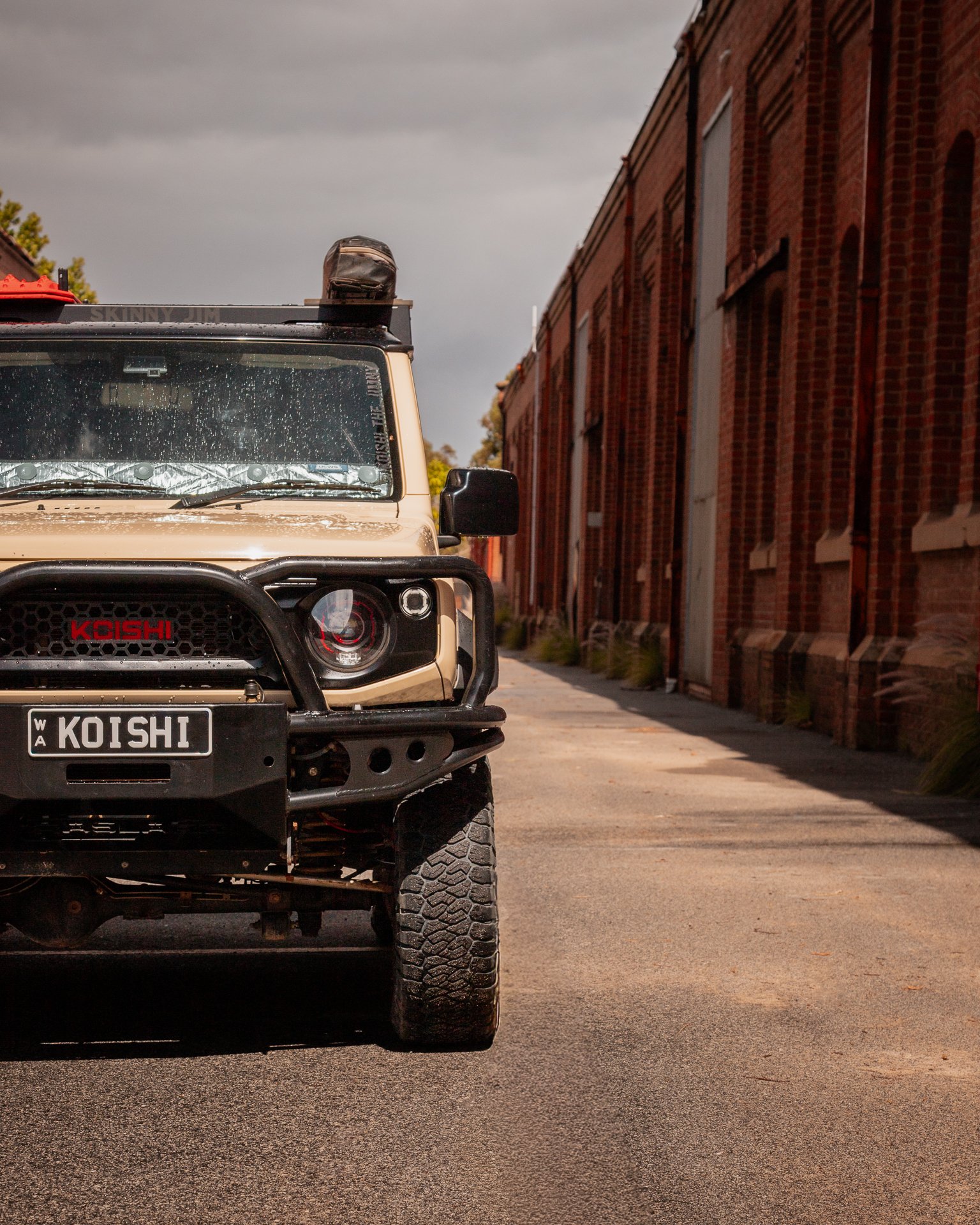 Front view of a beige JB74 Jimny with a Raslarr bull bar, parked on a street with red brick buildings on the right side, overcast sky in the background.