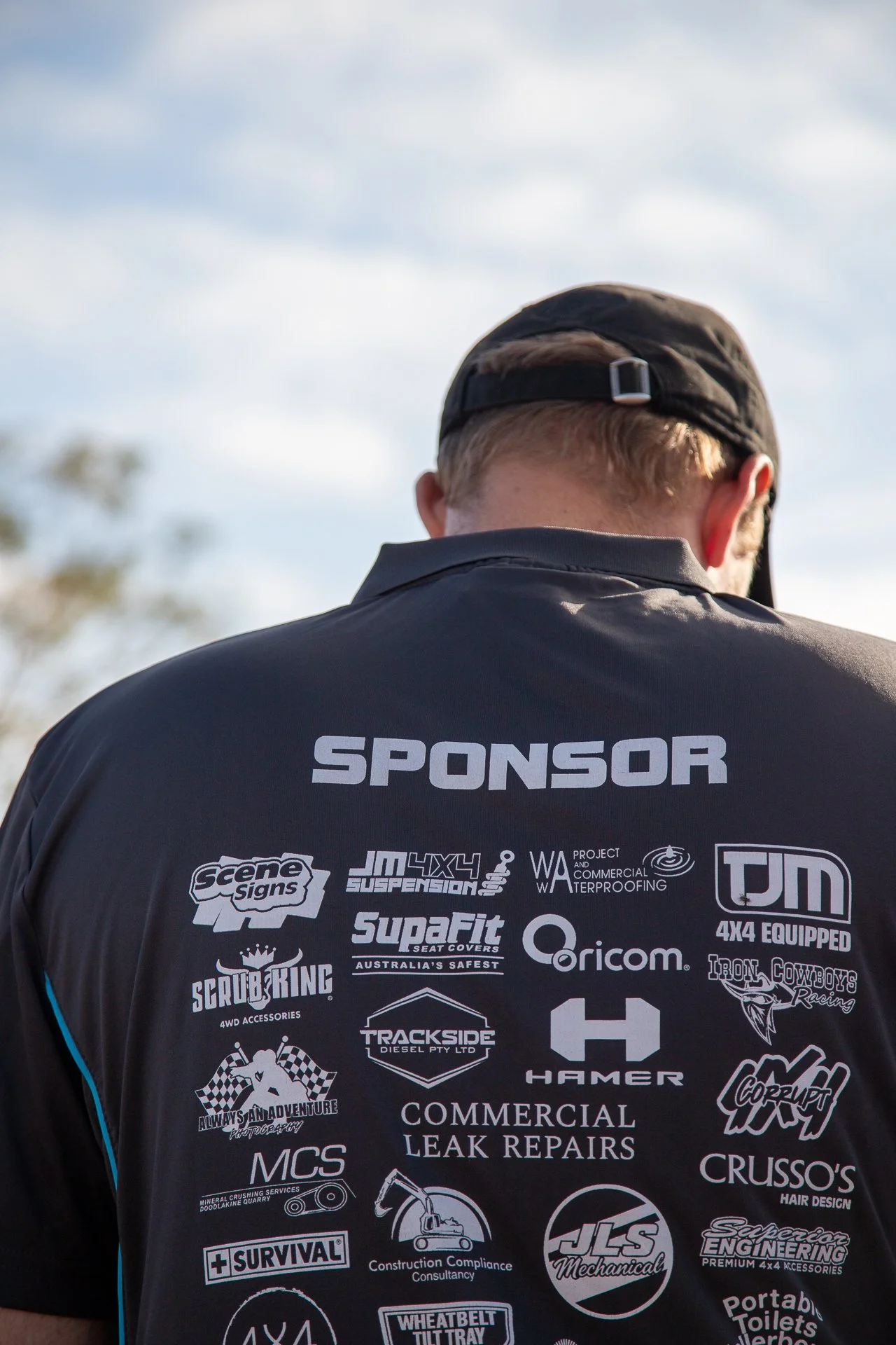 Back of a person wearing a black sponsorship shirt with logos of various off-road and automotive brands.