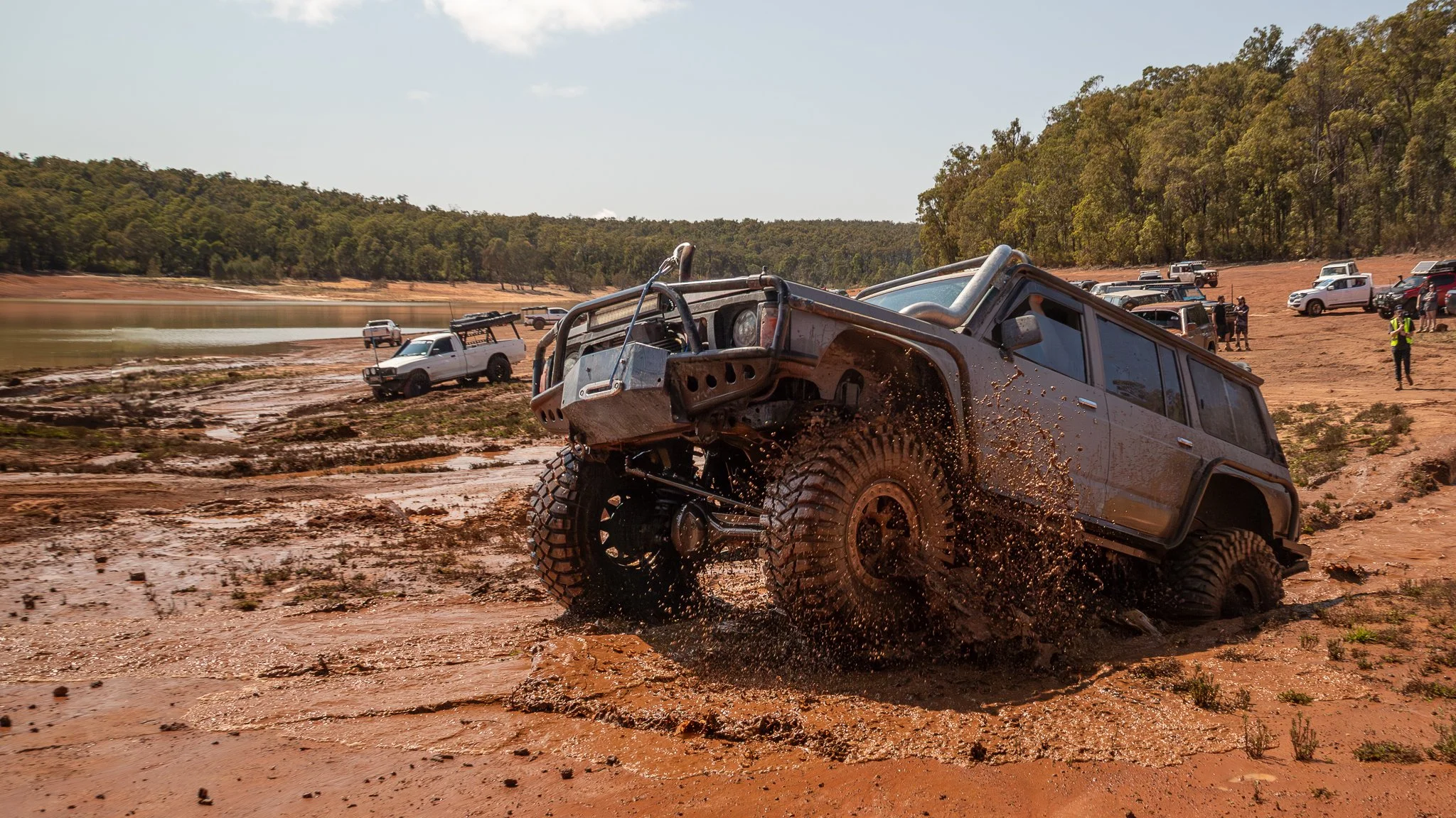 A rugged off-road vehicle splashing through muddy terrain near a body of water, with other vehicles and people in the background.