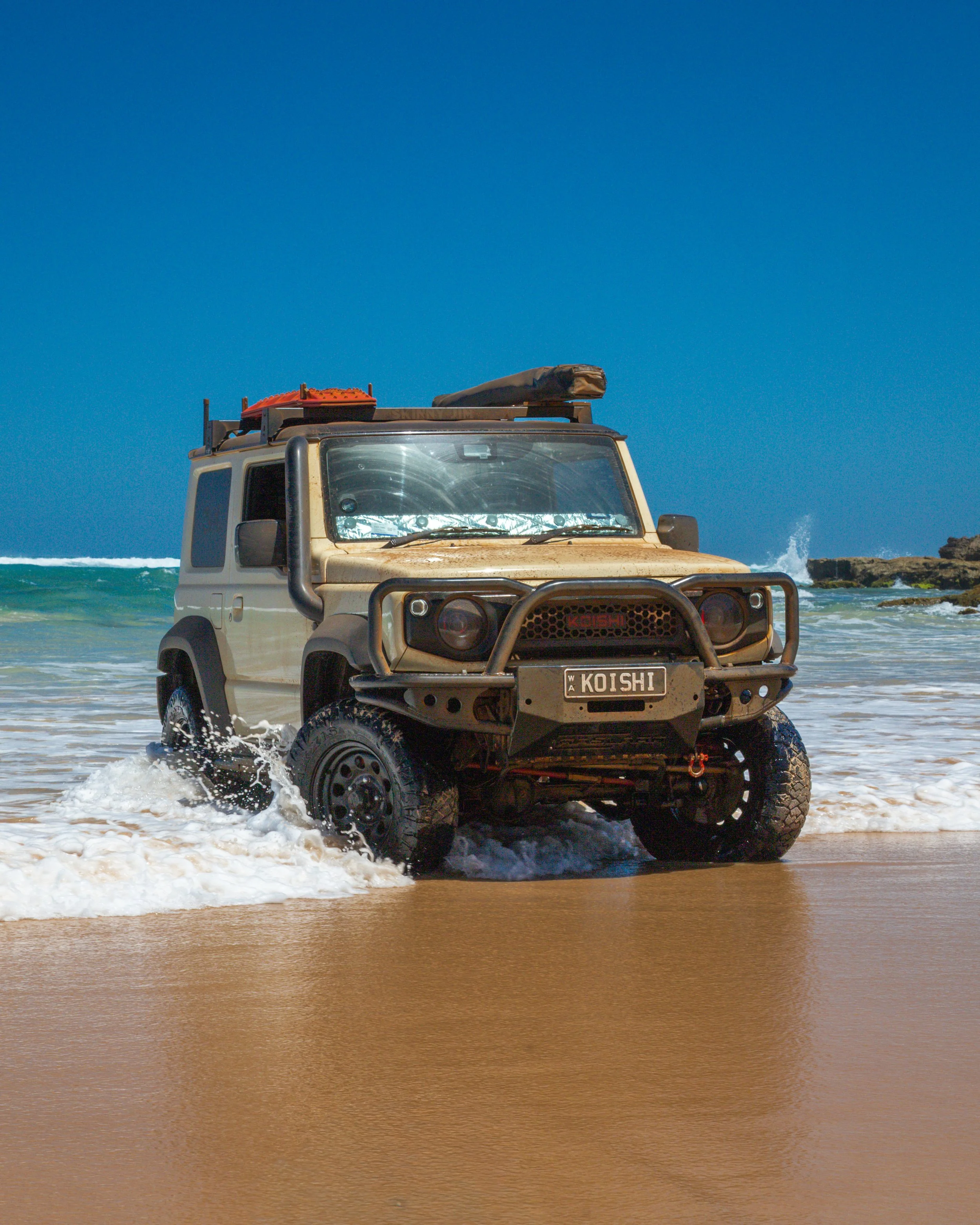 A beige off-road vehicle driving through shallow ocean water on a sandy beach, with a clear blue sky and rocks in the background.