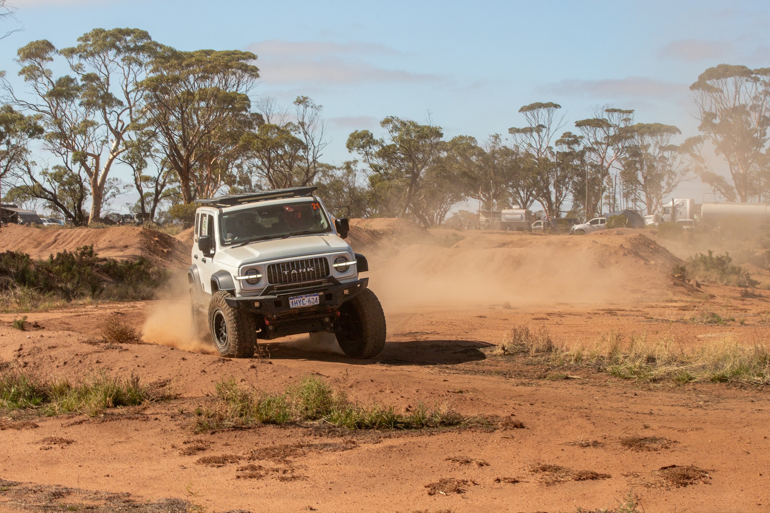 A white off-road vehicle kicking up dust while driving on a dirt track with trees and other vehicles in the background during daytime.